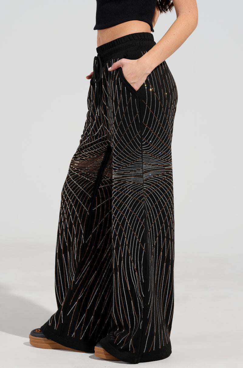 A person is wearing the "LIGHTS, CAMERA, ACTION! EMBELLISHED WIDE LEG SWEAT PANT," which comes in black with gold and silver geometric line patterns. These pants feature a drawstring waist, side pockets, and a high-rise waistband. They are styled with a sleek black top and matching footwear against a light-colored backdrop.