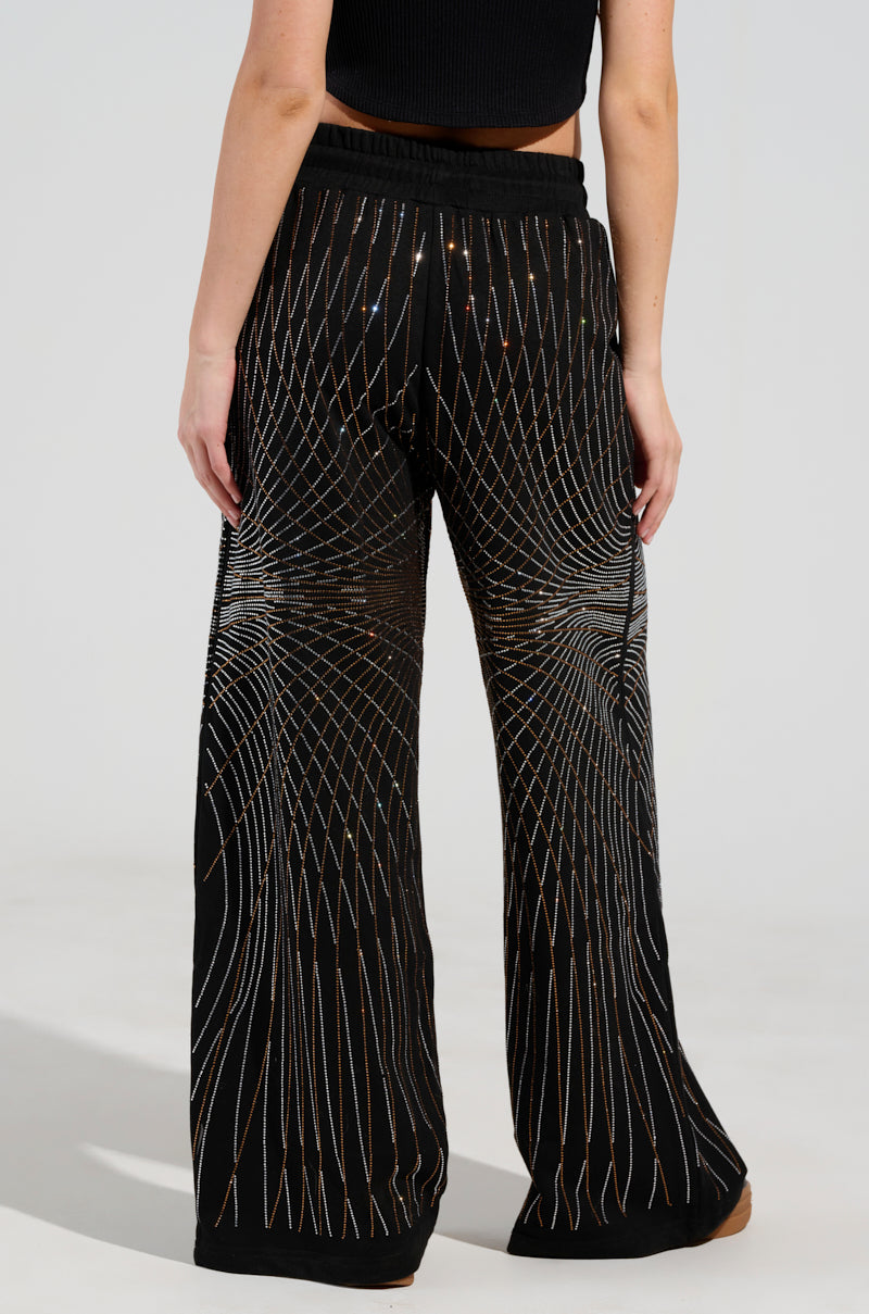 A person is seen from behind wearing the "LIGHTS, CAMERA, ACTION! EMBELLISHED WIDE LEG SWEAT PANT," which features a black design with a white geometric line pattern and a high-rise waistband. They are also styled in a black sleeveless top against a neutral background.