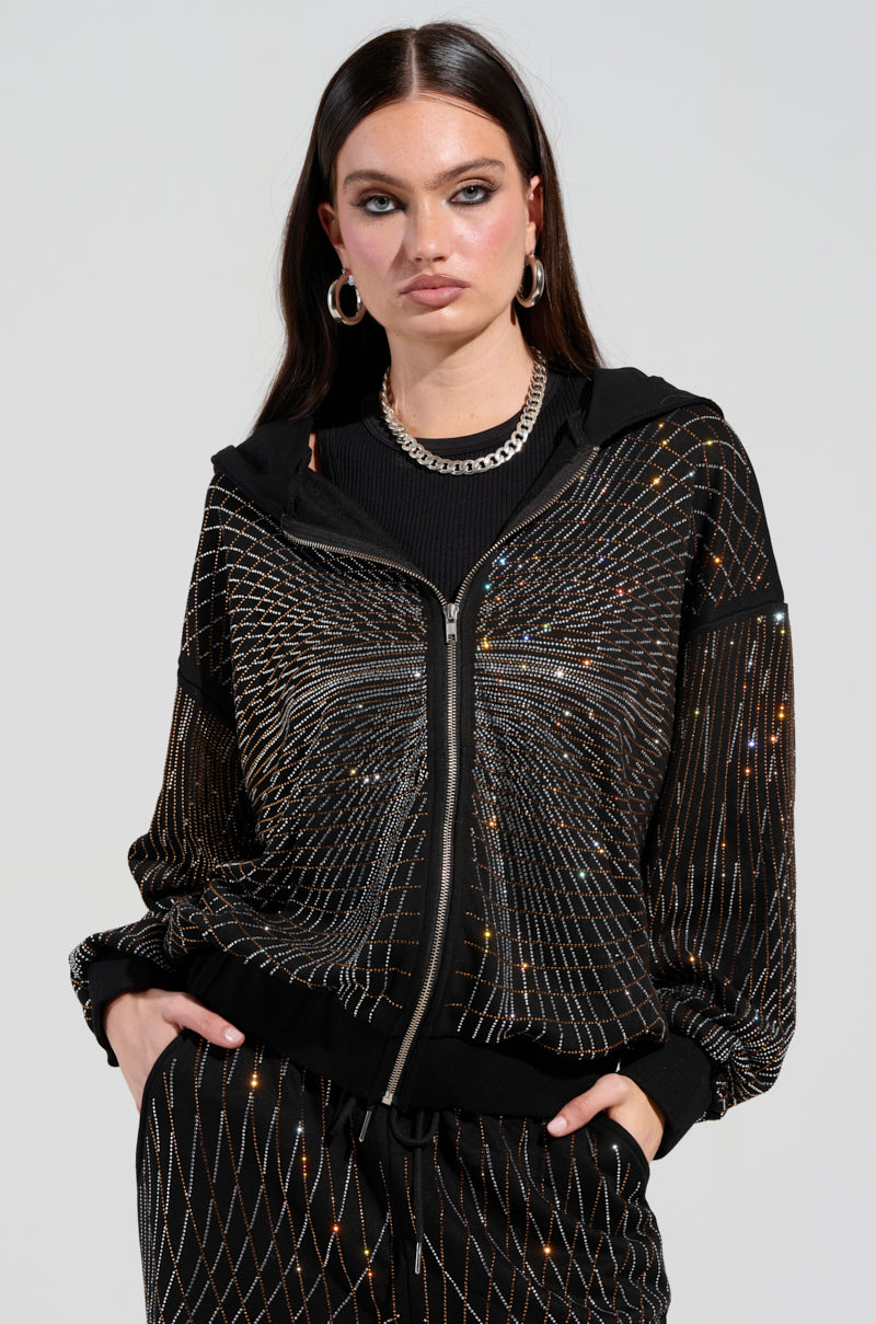 A person wearing the LIGHTS, CAMERA, ACTION! EMBELLISHED ZIP UP SWEATSHIRT paired with matching sparkling black pants stands with hands in pockets. The outfit showcases rhinestone embellishments and a geometric pattern with shimmering accents. They have long dark hair, hoop earrings, and a silver chain necklace against a plain background.