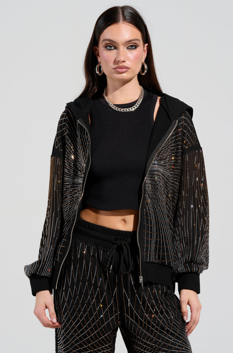 A person with long dark hair is wearing an oversized, shimmering black LIGHTS, CAMERA, ACTION! EMBELLISHED ZIP UP SWEATSHIRT over a cropped top and matching pants. They accessorize with hoop earrings and a chain necklace, while rhinestone embellishments catch the light against a plain background.