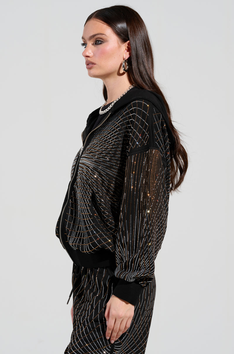 A woman with long dark hair is wearing the oversized LIGHTS, CAMERA, ACTION! Embellished Zip Up Sweatshirt in black, adorned with metallic geometric patterns. Rhinestone embellishments catch the light as she stands in profile against a plain background.