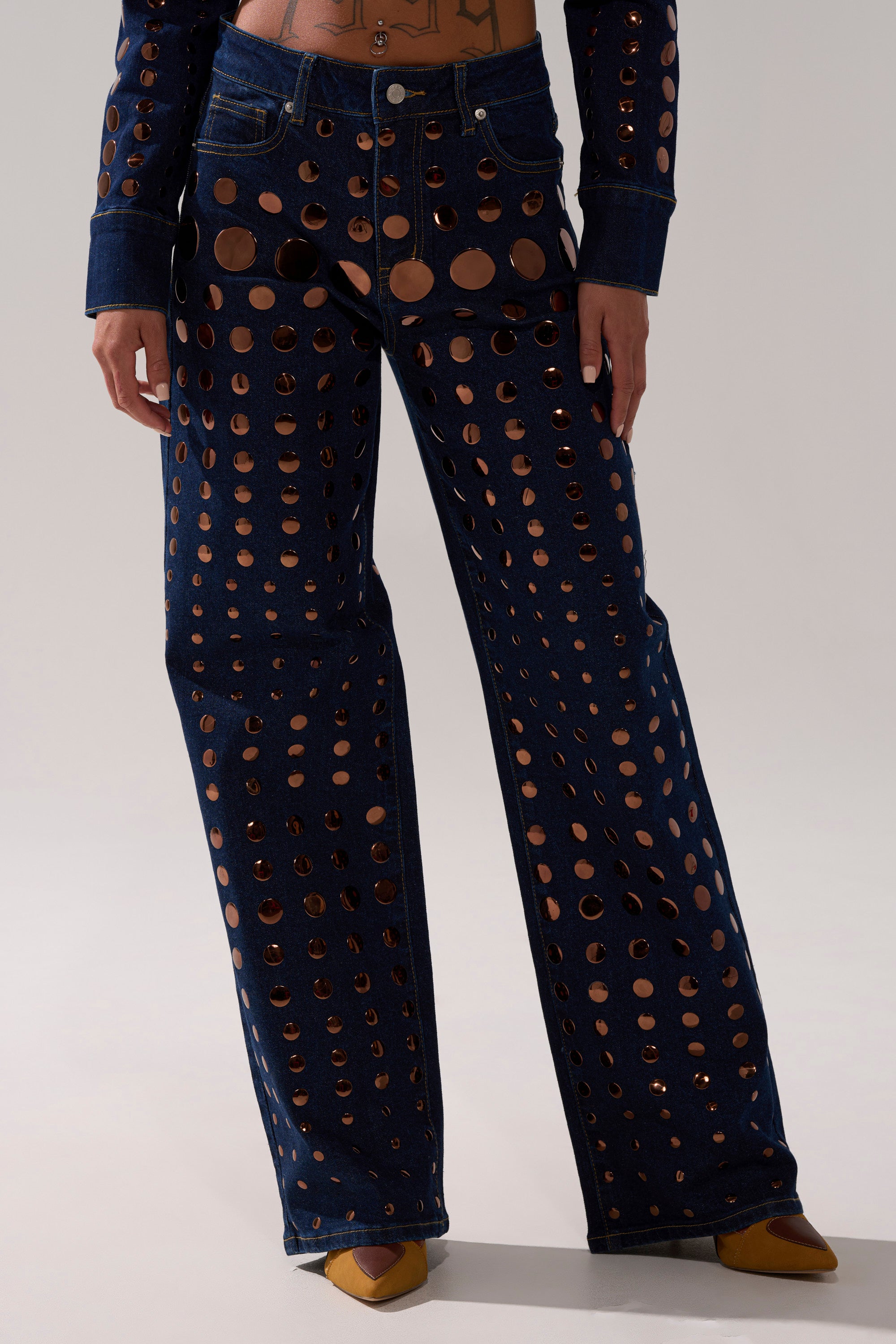 A person wears DISCO BABE RELAXED DENIM—high-waisted, wide-leg jeans with bold circular cutouts and metallic gold details. They have a tattooed torso, matching top, brown pointed shoes, and stand against a plain light background.