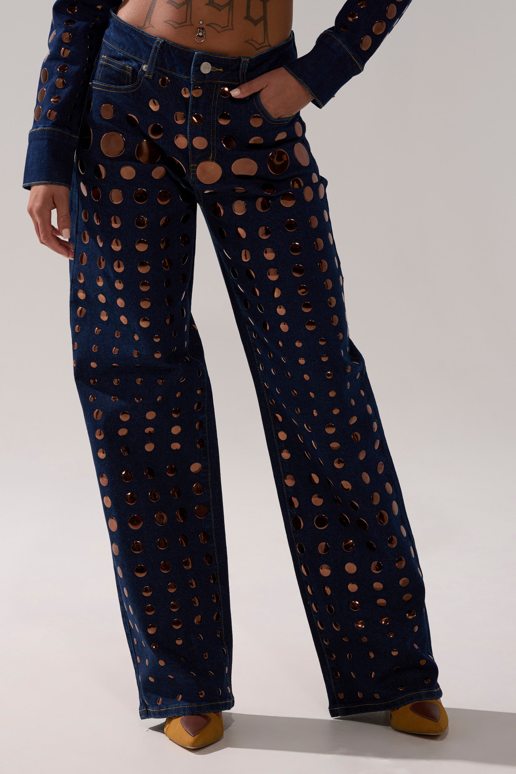 A person wearing the DISCO BABE RELAXED DENIM—high-waisted, wide-leg jeans with circular cutouts, gold embellishments—styled with a matching cropped top and yellow pointed-toe shoes.