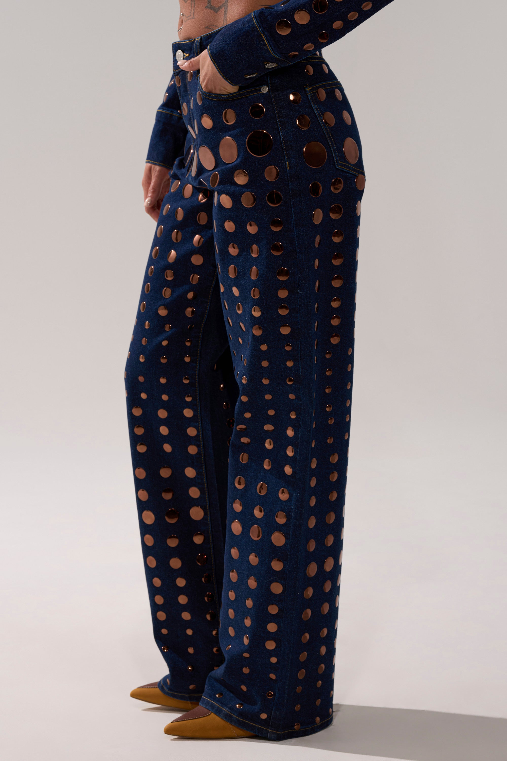 A person models the DISCO BABE RELAXED DENIM, high-waisted wide-leg jeans featuring large circular cutouts, paired with pointed brown shoes against a plain light background.
