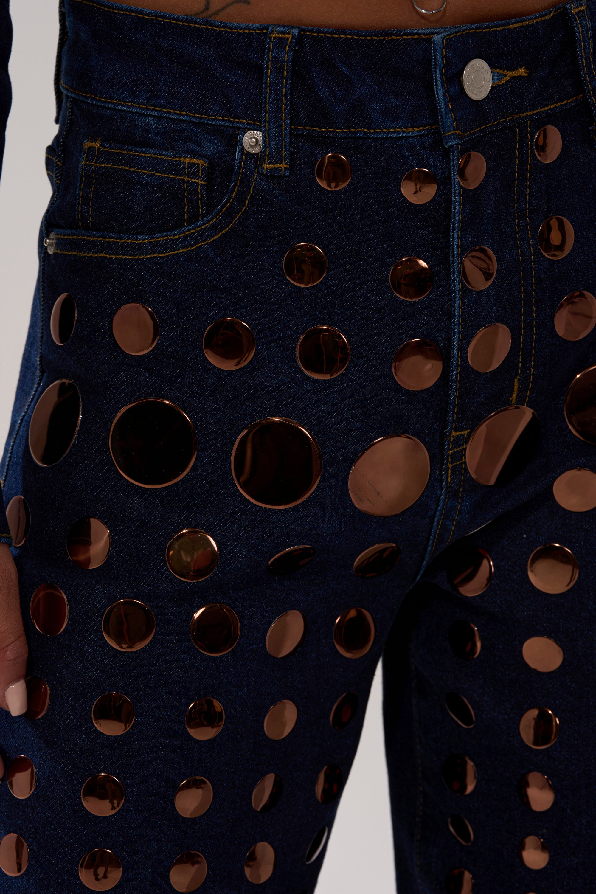 Close-up of the DISCO BABE RELAXED DENIM: high-waisted, wide-leg jeans in a dark wash with large and small gold metallic circular embellishments. A manicured hand rests at the side; a partial midriff appears at the top.