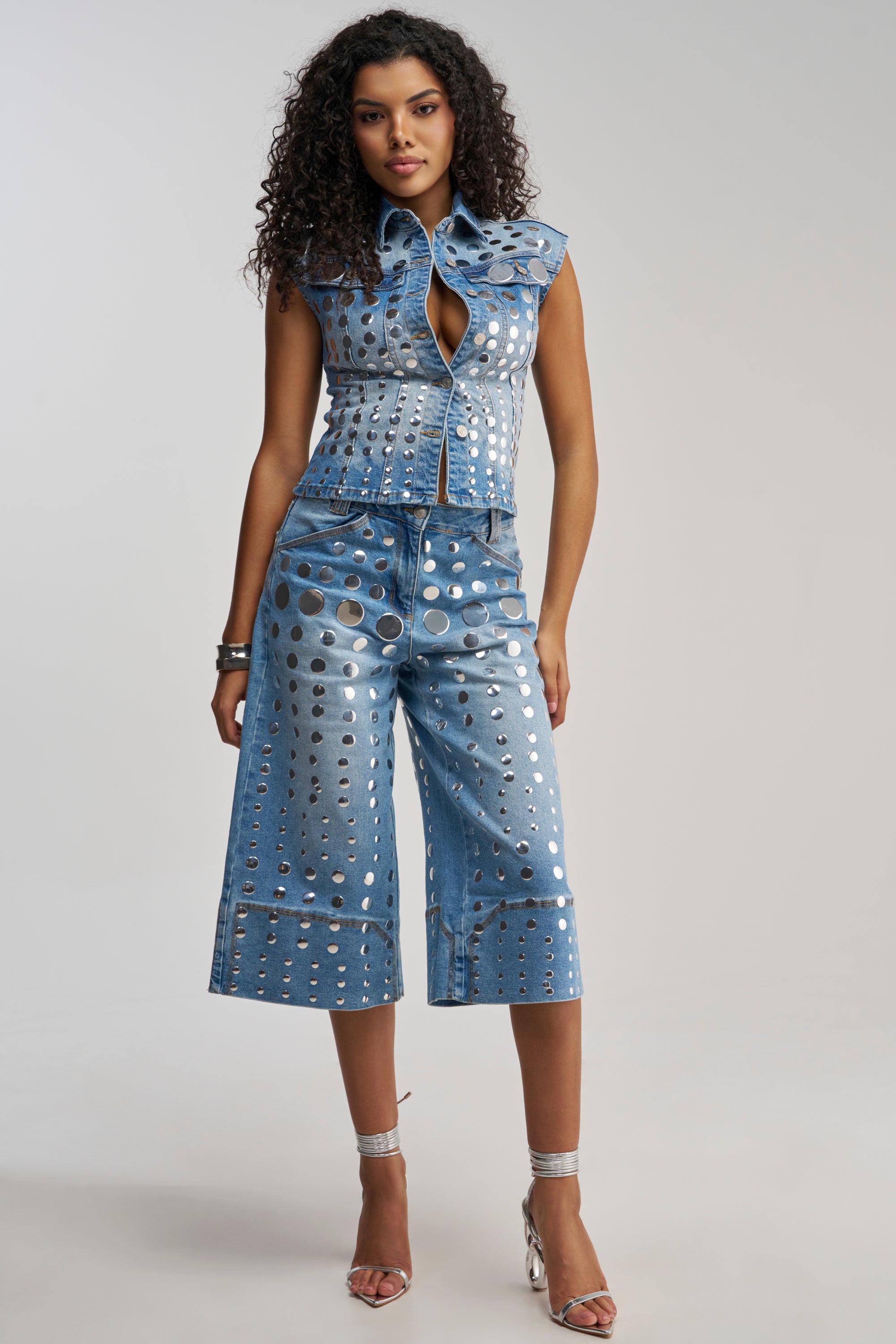 A woman with curly hair models the STUDS EVERYWHERE CULOTTE—a sleeveless, metallic-studded denim vest and matching culottes embellished with silver circles—paired with silver high heels, standing against a plain background.