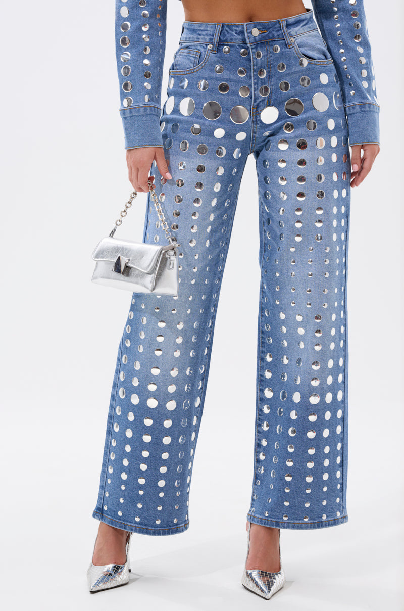 A person wears the STUDS EVERYWHERE RELAXED DENIM PANT with large studs and silver accents, paired with a matching top, a metallic silver handbag, and silver pointed high heels.