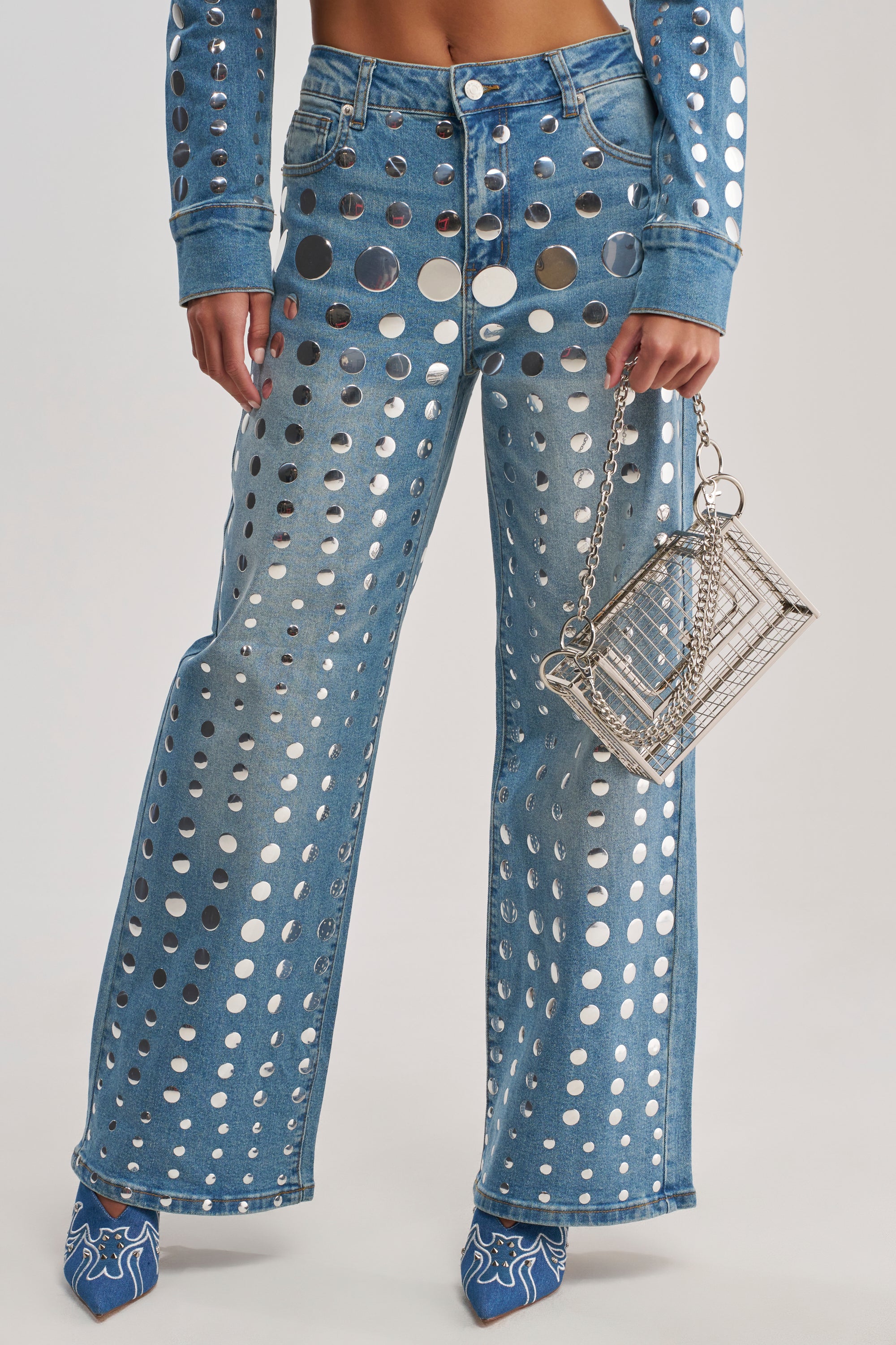 Wearing the STUDS EVERYWHERE RELAXED DENIM PANT and a matching jacket with bold silver grommet holes, a person carries a clear rectangular handbag with a chain strap and sports blue heeled shoes with white details.