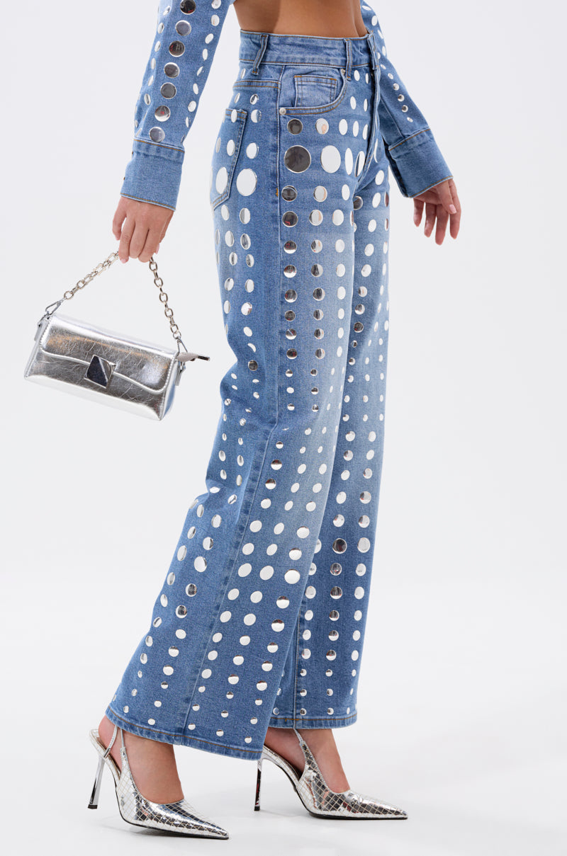 A person wears the STUDS EVERYWHERE RELAXED DENIM PANT with metallic circular cutouts, a matching denim jacket, silver pointed-toe heels, and carries a small metallic chain-strap handbag.