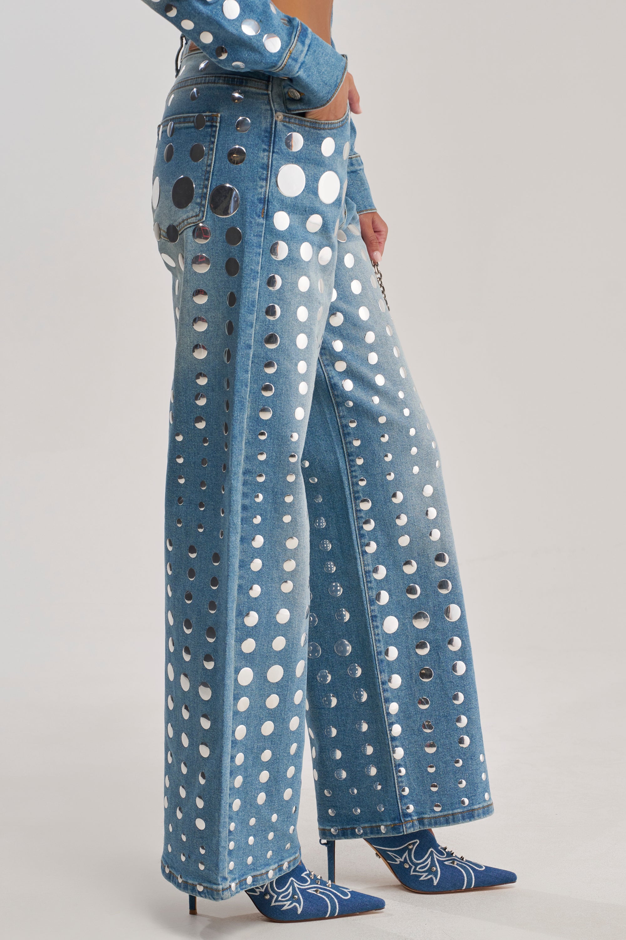 A person wears the STUDS EVERYWHERE RELAXED DENIM PANT, featuring a relaxed fit, wide-leg silhouette, and silver stud embellishments, paired with a matching top and pointed blue heels with a white pattern.