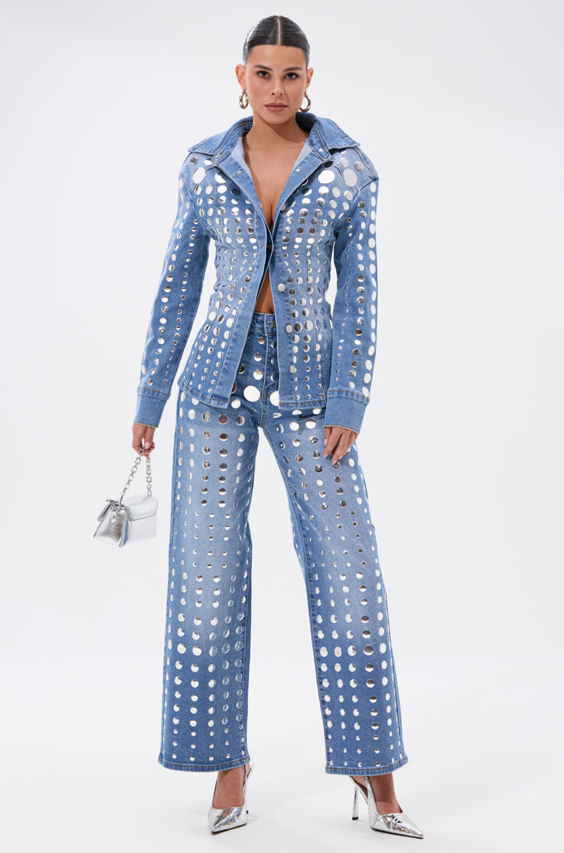 A woman stands confidently in the STUDS EVERYWHERE RELAXED DENIM PANT with matching high-rise jacket, both detailed with metallic circular cutouts. She accessorizes with a small silver handbag and pointed silver heels, her hair slicked back and center-parted.