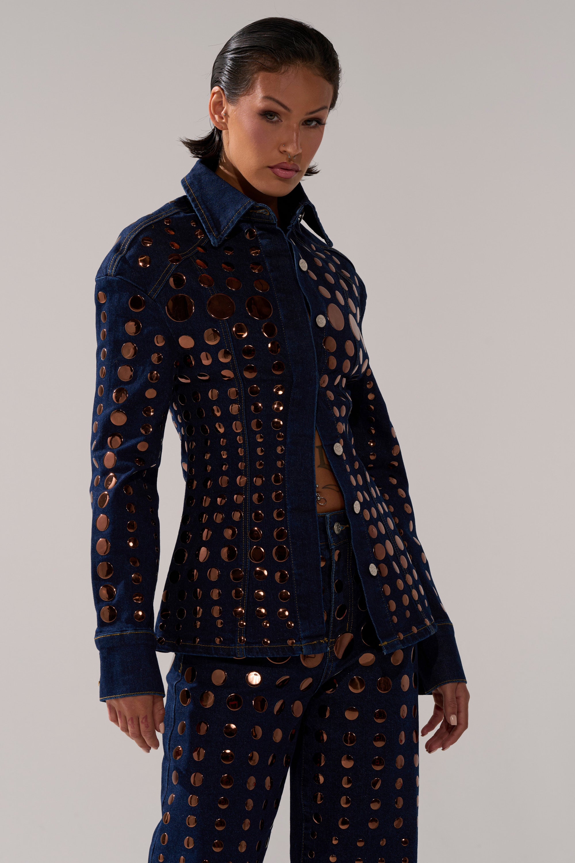 A woman confidently models the DISCO BABE DENIM BUTTON DOWN, a dark-wash denim top featuring circular cutouts and metallic accents, posing against a plain light background to showcase its bold, embellished style.
