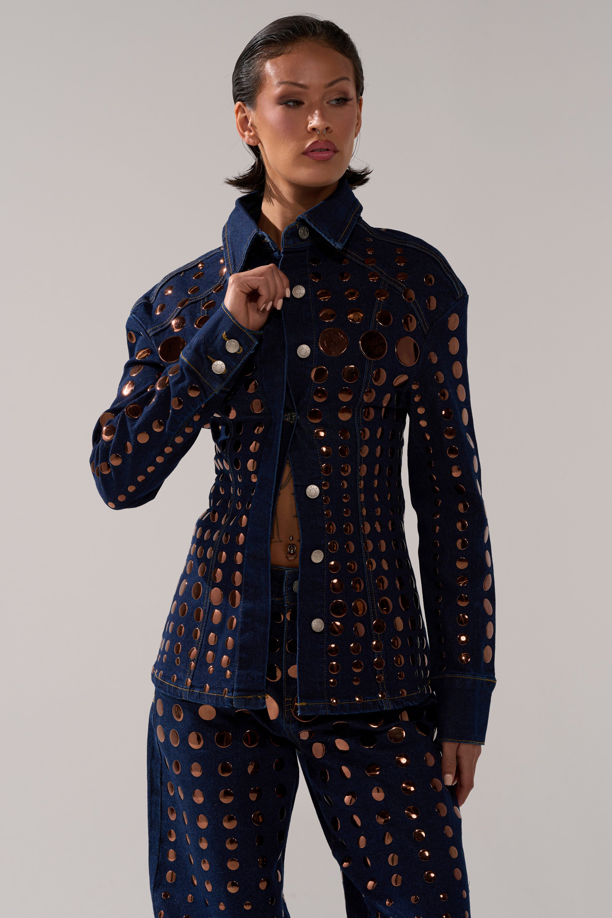 A woman poses confidently to the side in the DISCO BABE DENIM BUTTON DOWN, a dark-wash denim top with metal eyelet cutouts, featuring evenly spaced circular holes for a bold, modern look.