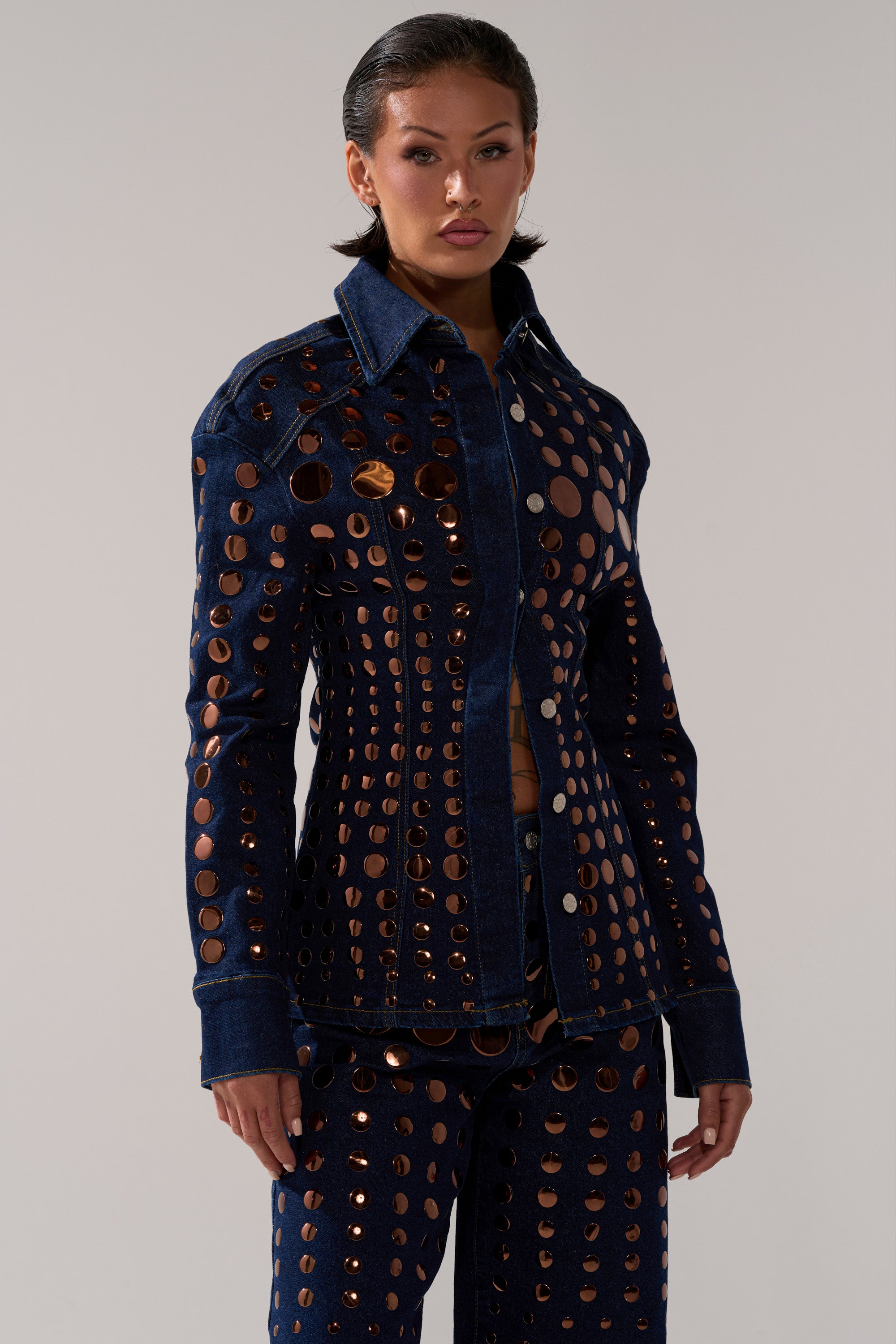 A woman wears the DISCO BABE DENIM BUTTON DOWN and matching pants with large circular cutouts revealing copper fabric underneath. She has slicked-back hair and poses against a plain light background.