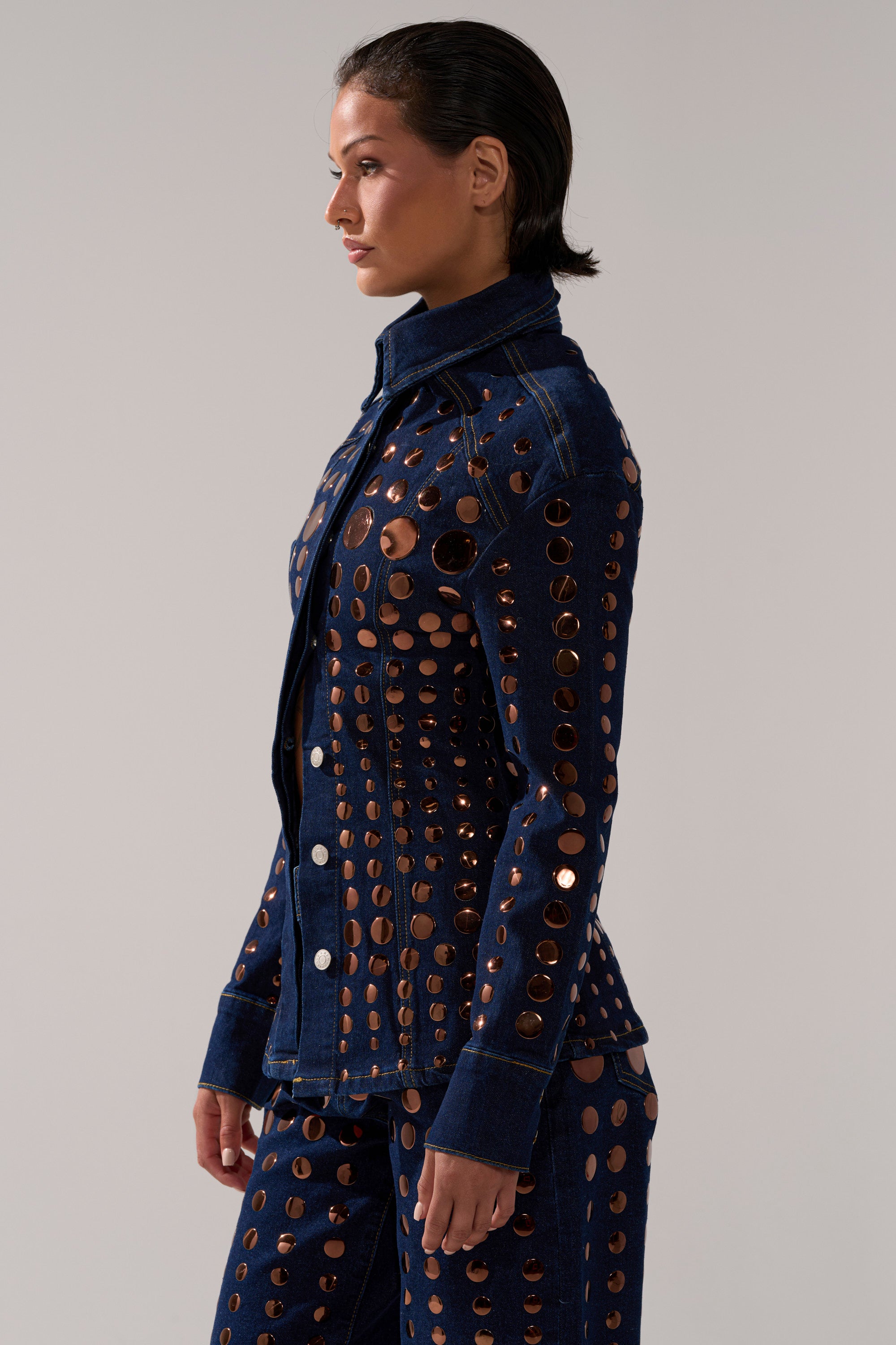 A woman with slicked-back dark hair wears a fitted DISCO BABE DENIM BUTTON DOWN featuring circular cut-out holes, standing in profile against a plain light background.