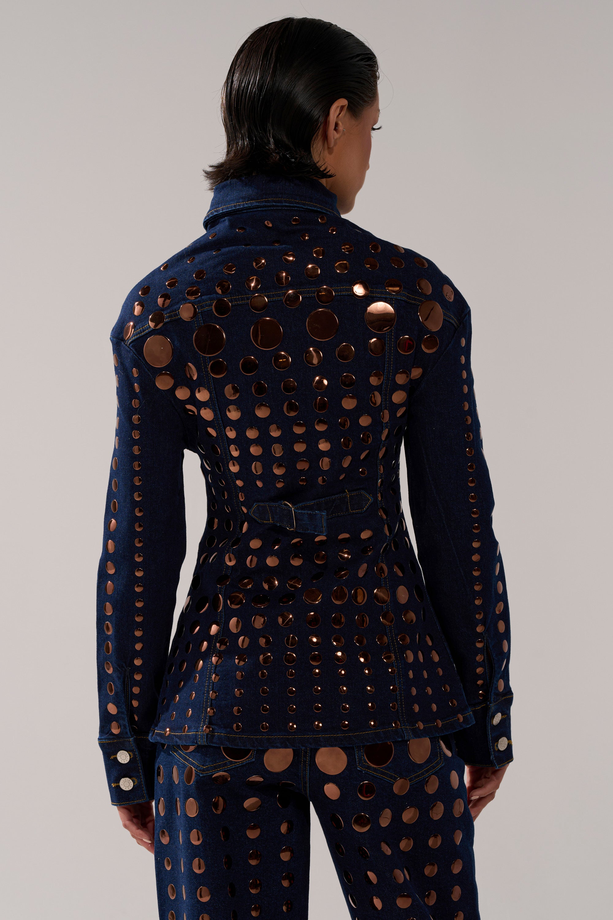 Seen from behind, a person with short dark hair wears the DISCO BABE DENIM BUTTON DOWN and matching pants covered in shiny copper studs and cutouts, standing against a plain light gray background.