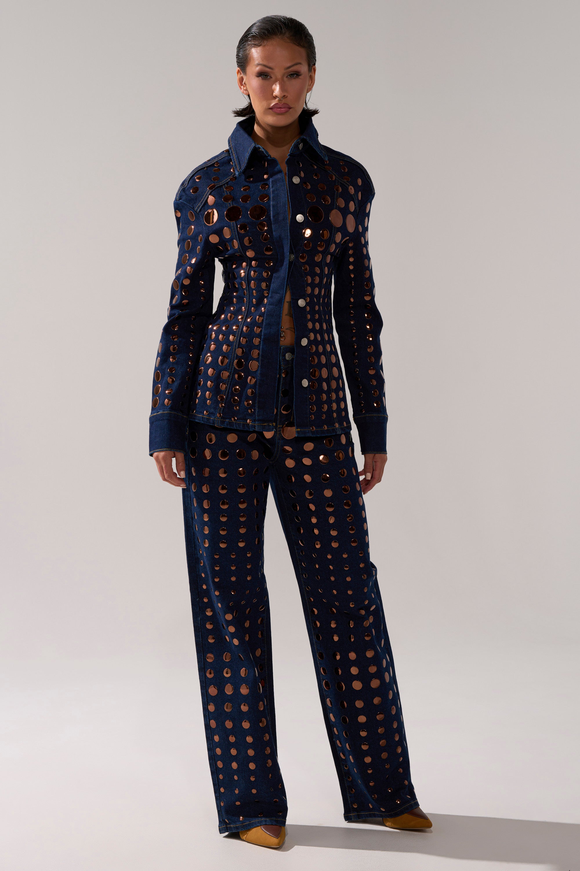 A woman wears the DISCO BABE DENIM BUTTON DOWN in a matching dark-wash set with circular cutouts, her hair slicked back as she poses against a plain light background.