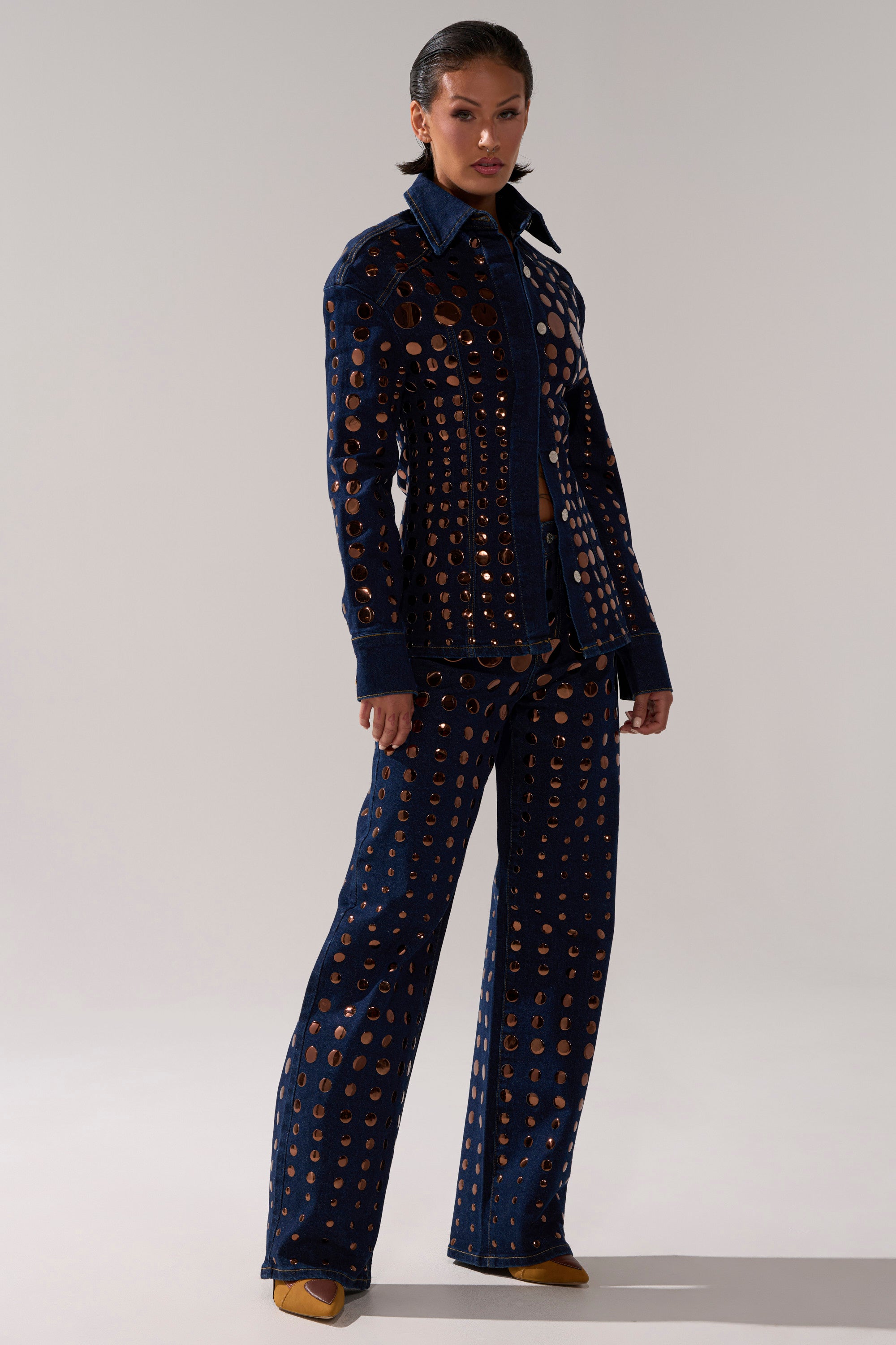 A woman exudes confidence in the DISCO BABE DENIM BUTTON DOWN, styled with wide-leg, dark-wash pants featuring metallic grommet details. Her slicked-back hair and the neutral background highlight the bold denim look.