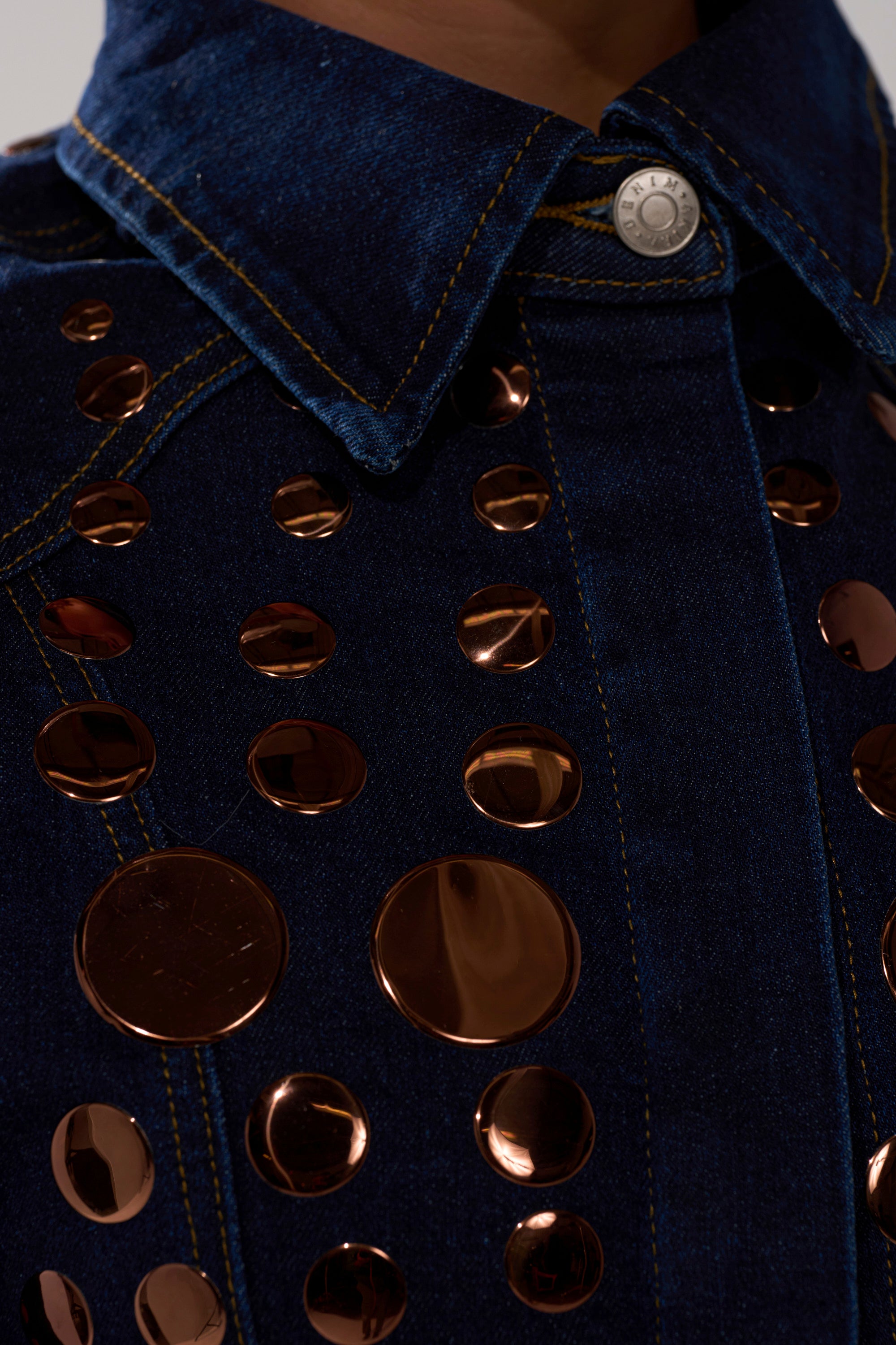 Close-up of the DISCO BABE DENIM BUTTON DOWN in dark blue, detailed with large copper metallic studs, gold-tone accents, yellow stitching, and a silver snap button at the collar.