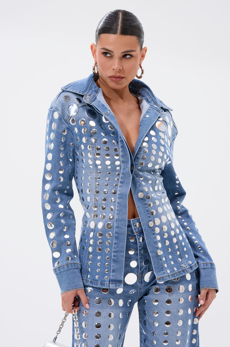 A woman wears the STUDS EVERYWHERE BUTTON DOWN denim jacket and matching pants with large silver grommets, slicked-back hair, hoop earrings, and a small silver bag, posing against a plain white background.