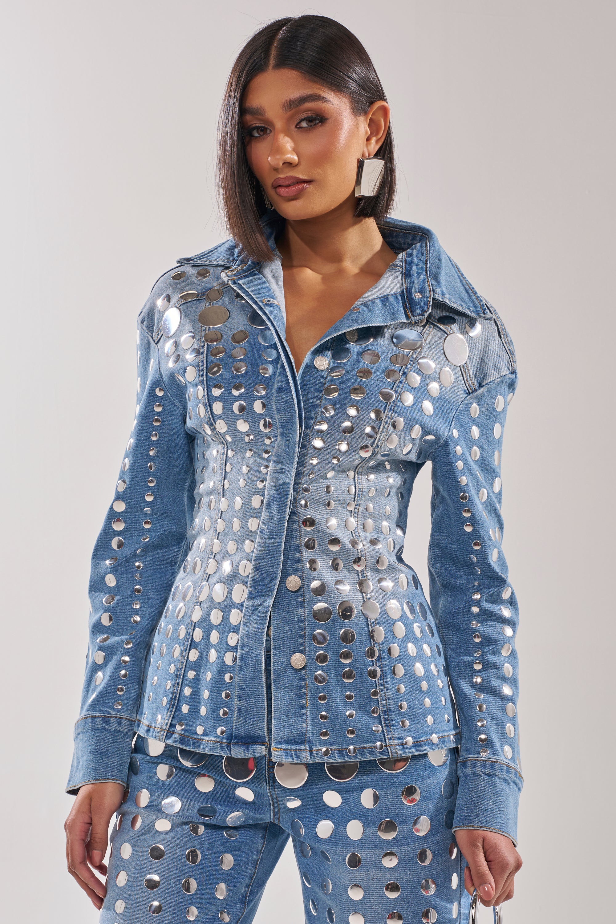 A woman wears the STUDS EVERYWHERE BUTTON DOWN and matching pants with metallic cutouts. Her hair is sleek and straight, she sports large square earrings, and poses against a plain light background. 0