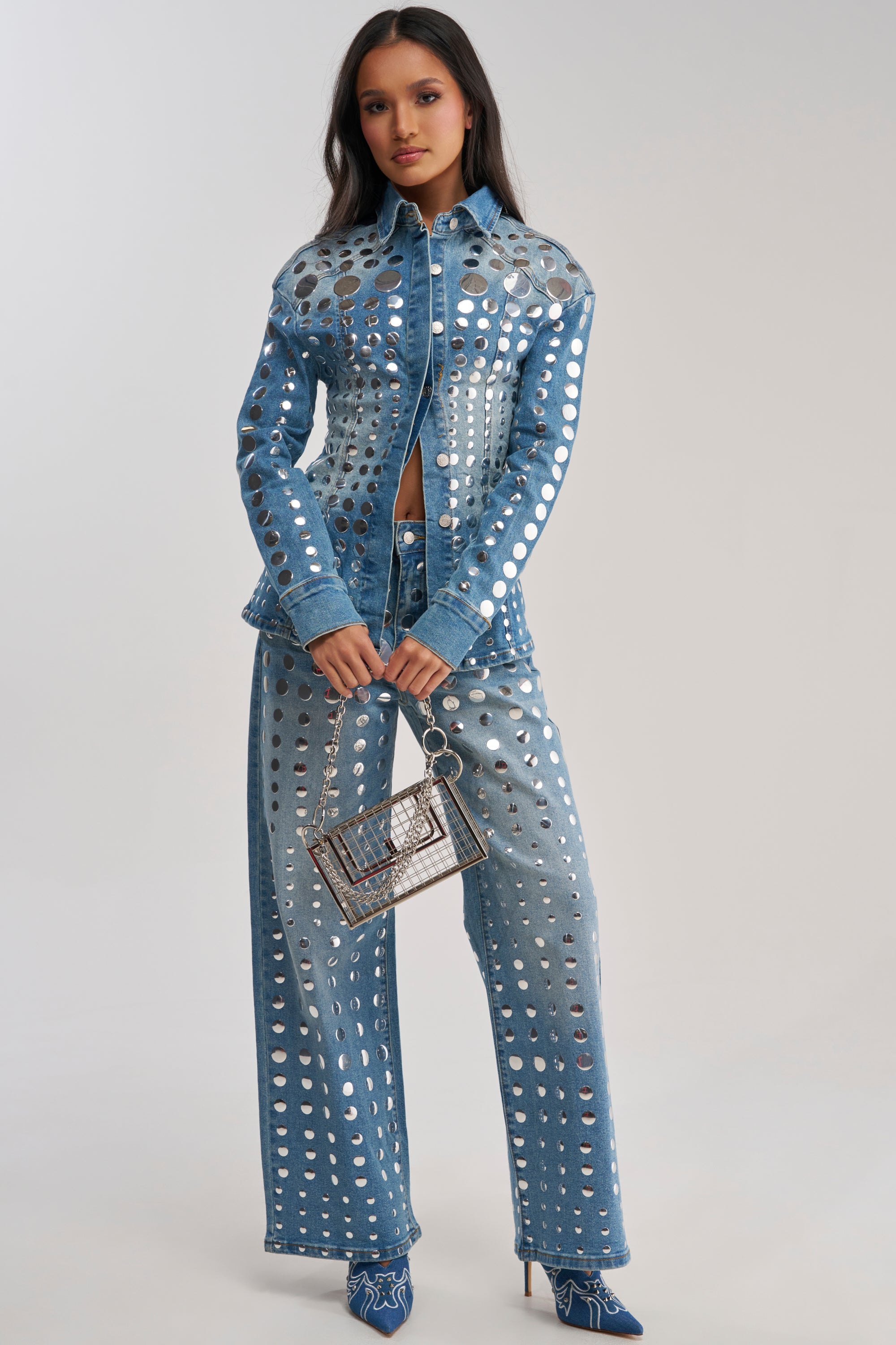 A woman faces forward, wearing the STUDS EVERYWHERE BUTTON DOWN denim top and matching pants set with large circular cutouts, a small transparent handbag, and blue high-heeled boots.