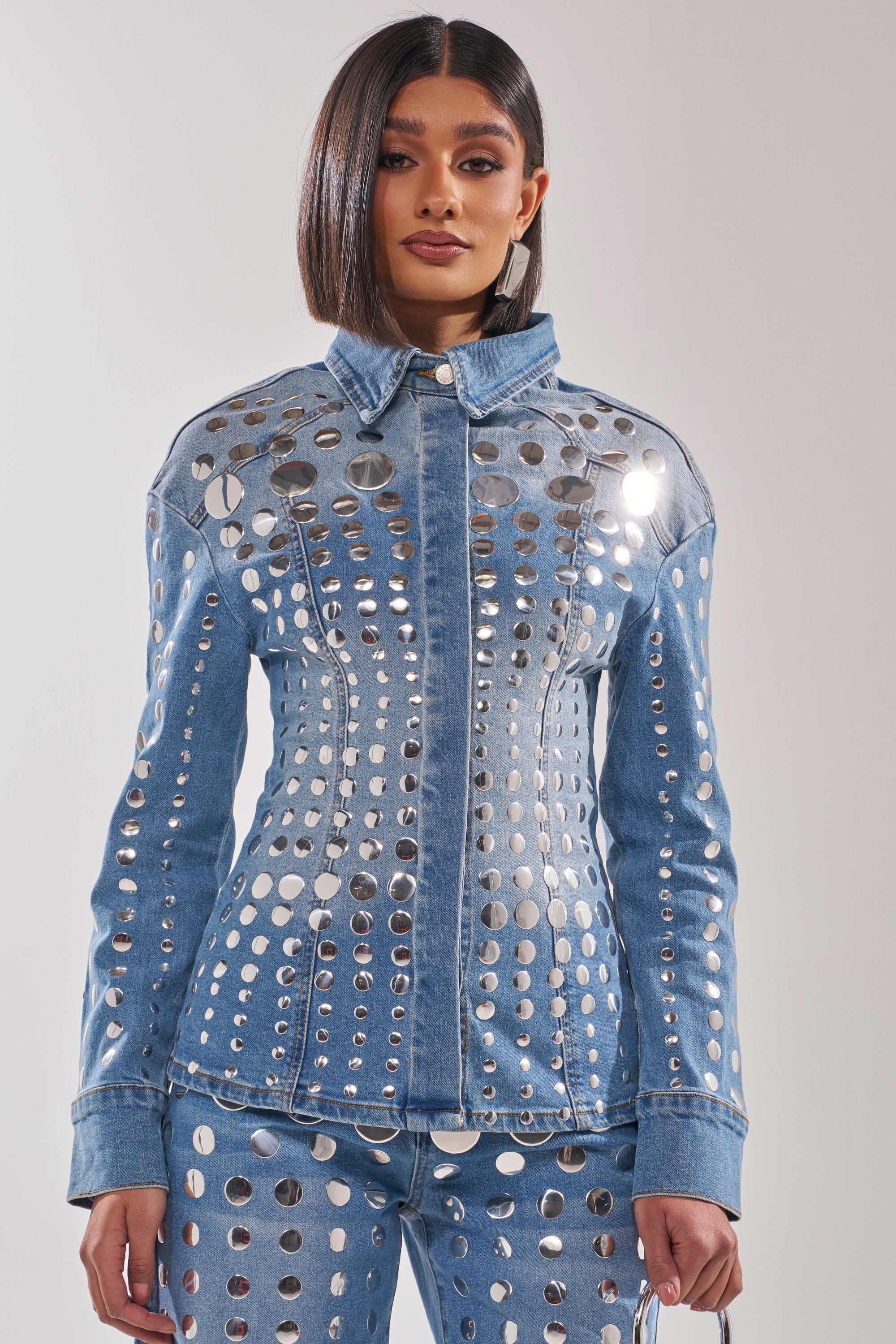 A woman with straight, shoulder-length hair models the STUDS EVERYWHERE BUTTON DOWN jacket and matching pants, both featuring metallic circular cutouts and a waist-contouring fit, against a plain light background. 1