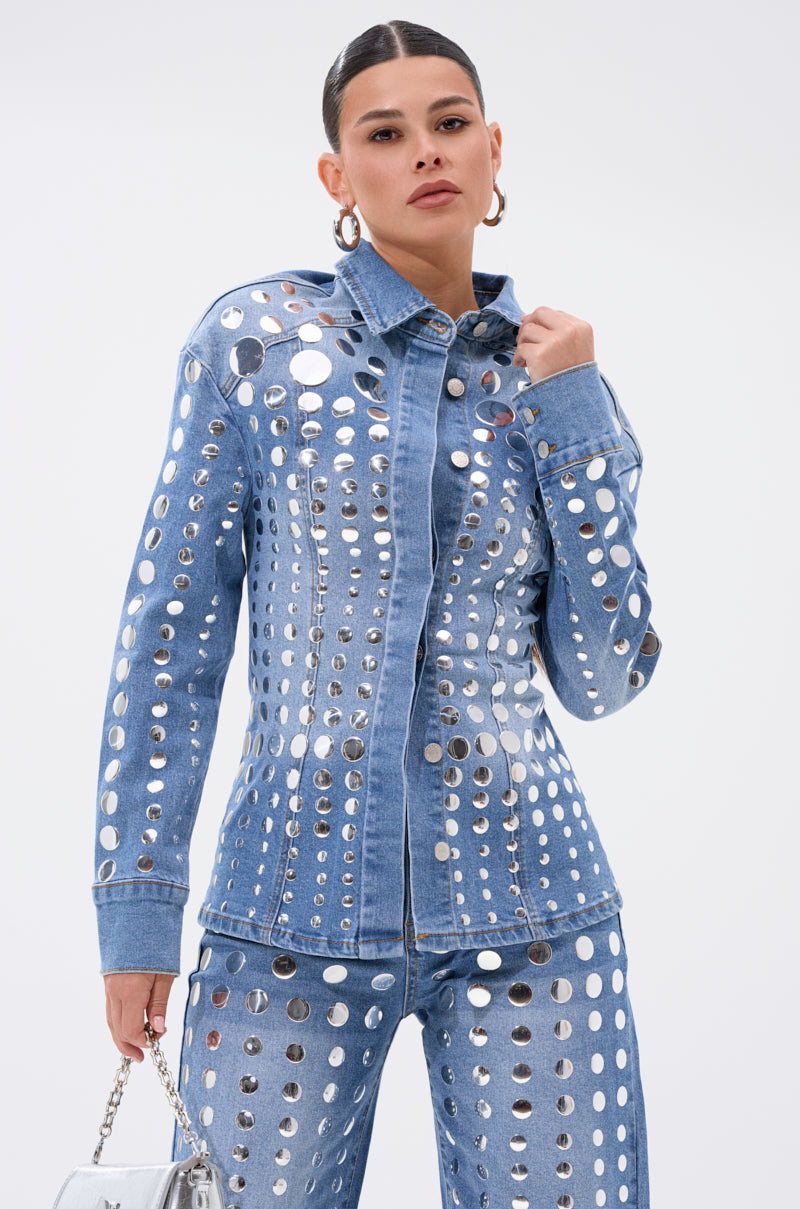 A woman stands confidently against a plain white background, wearing blue denim jeans with metallic cutouts and the STUDS EVERYWHERE BUTTON DOWN top. She completes the look with a silver handbag.
