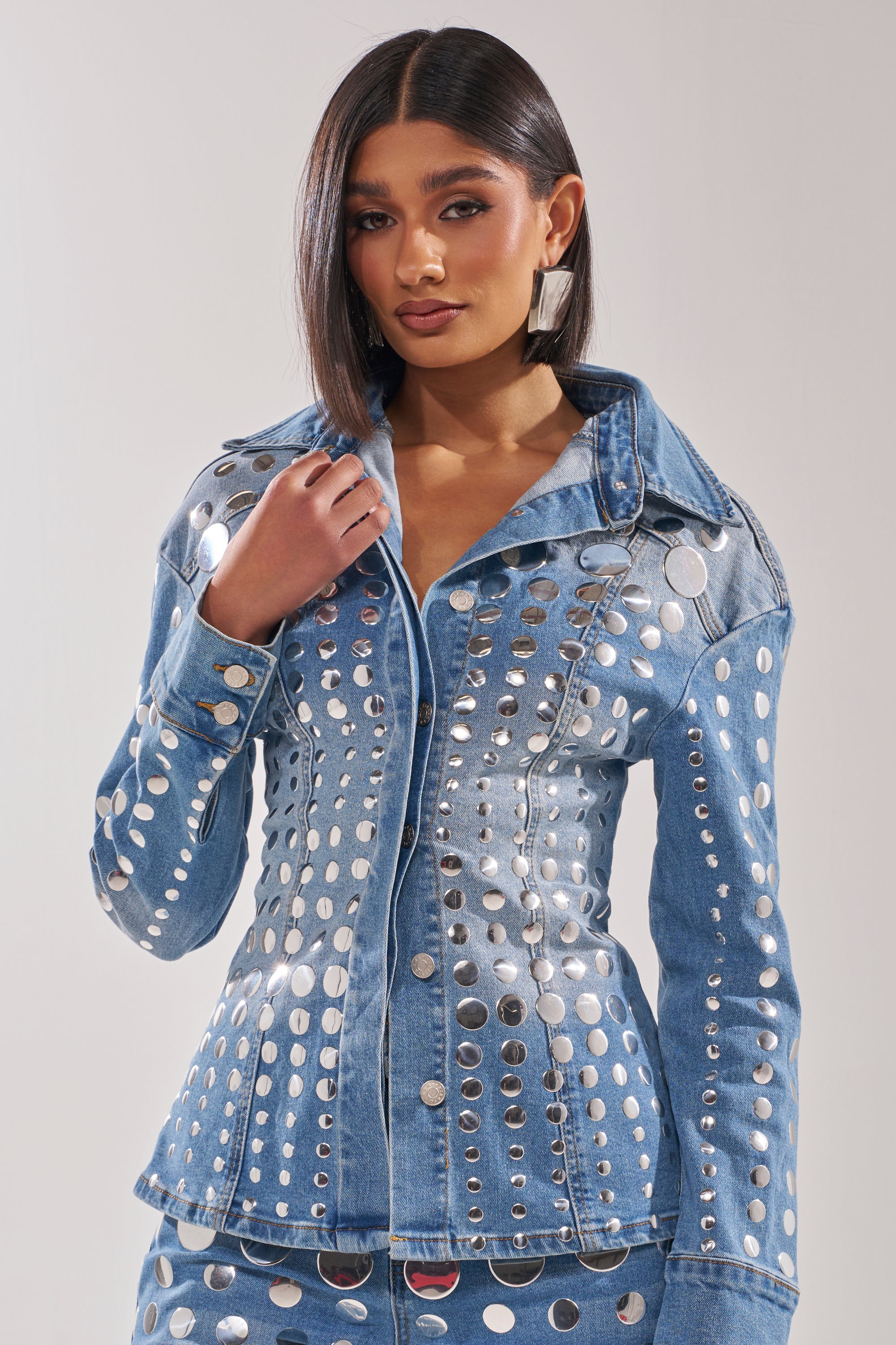 A woman with straight dark hair wears the STUDS EVERYWHERE BUTTON DOWN, a fitted denim top with studs and a waist-defining silhouette. She stands against a plain light background, gazing forward while touching the collar. 2
