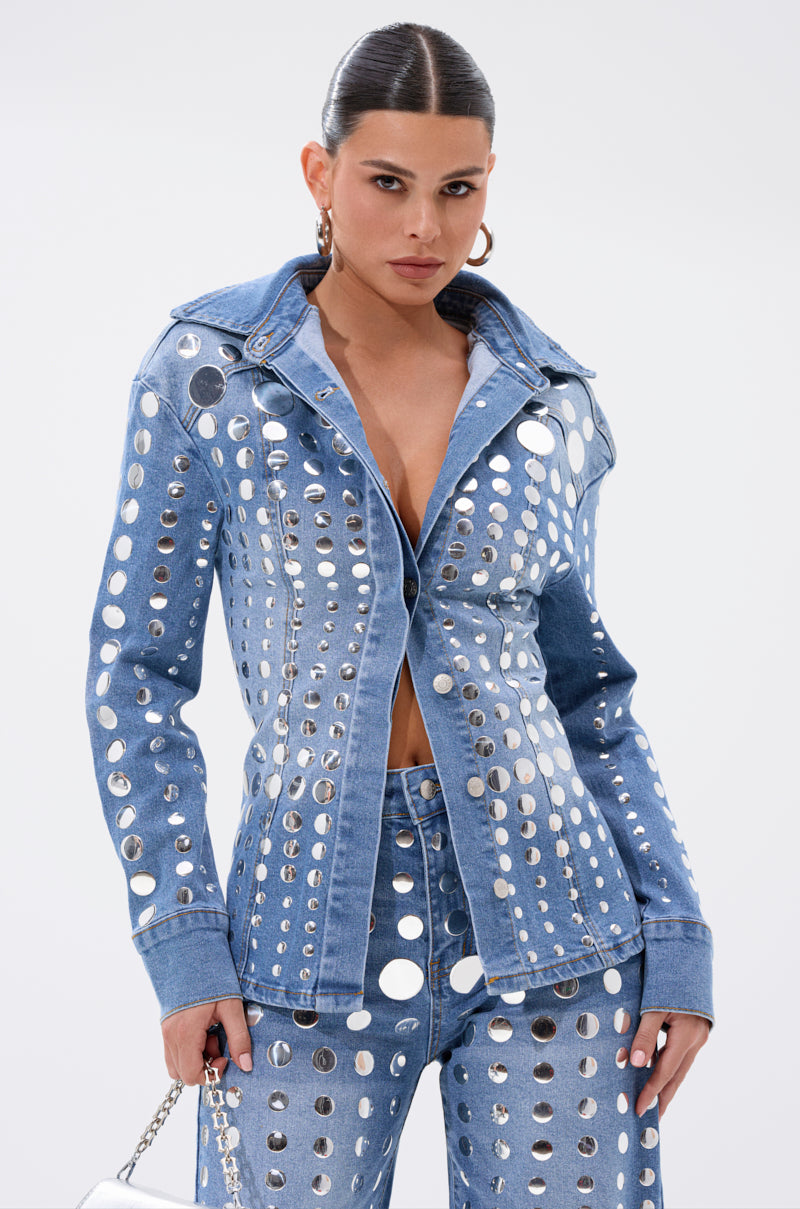 A woman with slicked-back hair wears the STUDS EVERYWHERE BUTTON DOWN, a blue denim jacket with large circular cutouts and a waist-contouring fit, paired with matching jeans. She poses confidently against a plain white background holding a small handbag.