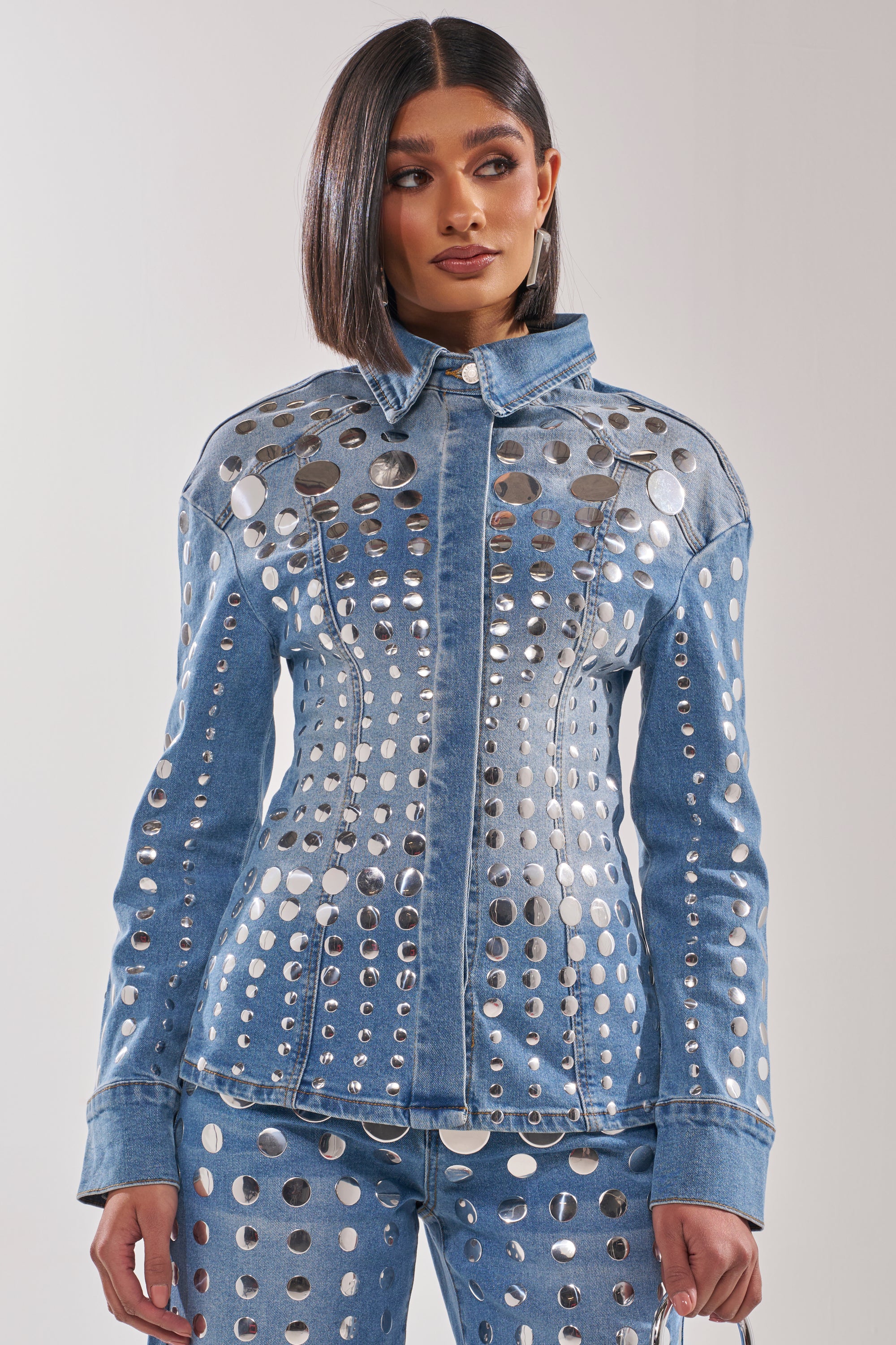 Wearing the STUDS EVERYWHERE BUTTON DOWN, a woman with straight, shoulder-length hair showcases its fitted, waist-contouring silhouette and bold rows of shiny metallic studs, paired with matching pants against a simple backdrop. 3