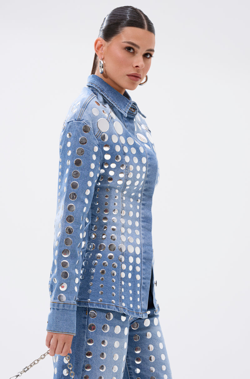 A woman with sleek, pulled-back hair poses sideways against a plain white background, wearing the STUDS EVERYWHERE BUTTON DOWN—a blue denim piece adorned with rows of large silver studs for a flattering shape.
