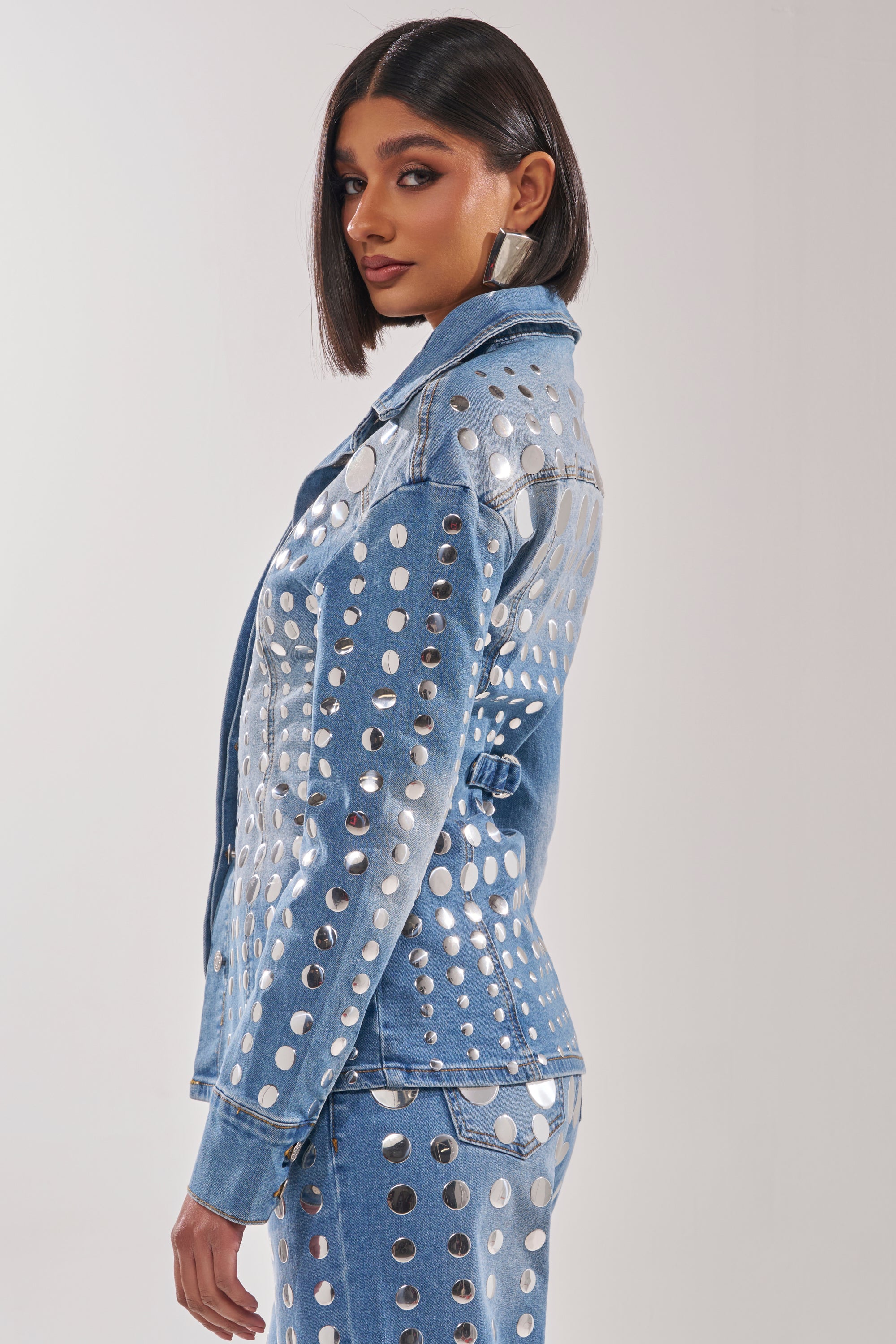 A woman with straight, chin-length dark hair wears the STUDS EVERYWHERE BUTTON DOWN—a studded denim top with a waist-contouring silhouette—as she stands sideways and looks over her shoulder. 4