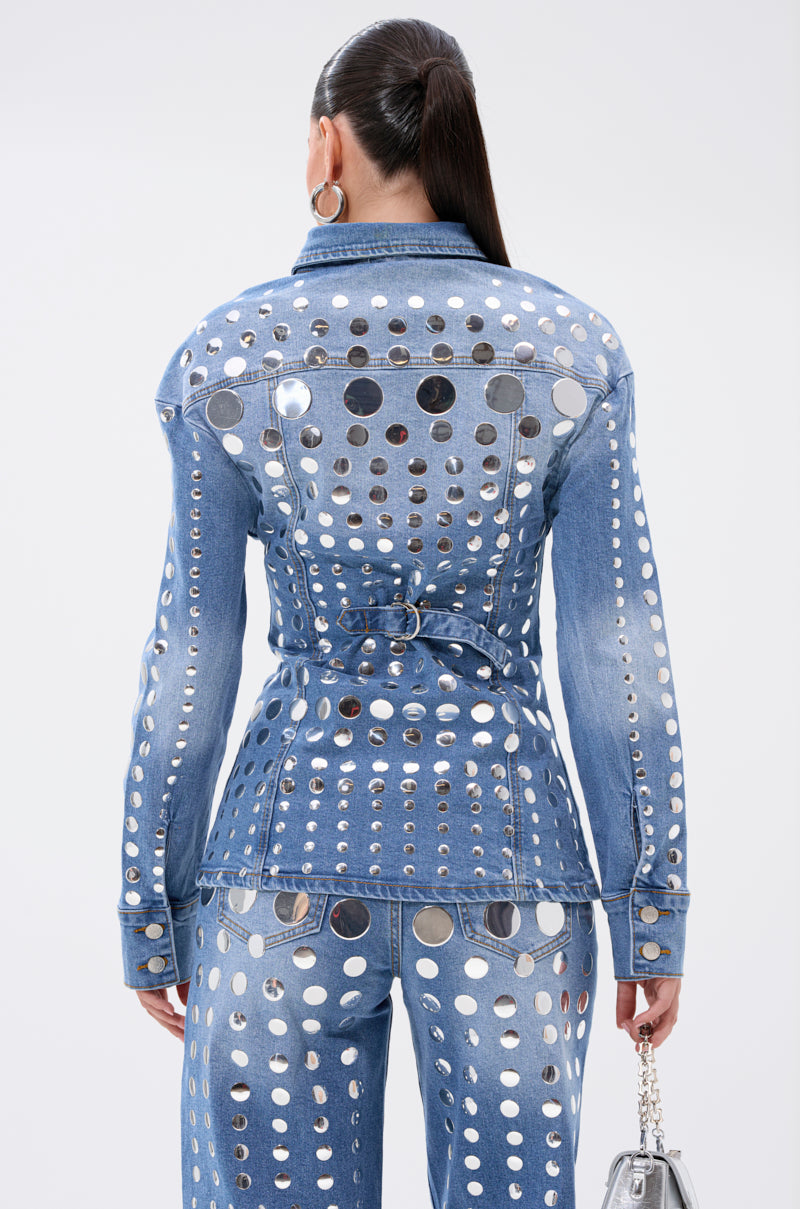 A woman with a ponytail is seen from the back, wearing the STUDS EVERYWHERE BUTTON DOWN denim top and jeans that contour her waist. She accessorizes with a silver handbag and large hoop earrings.
