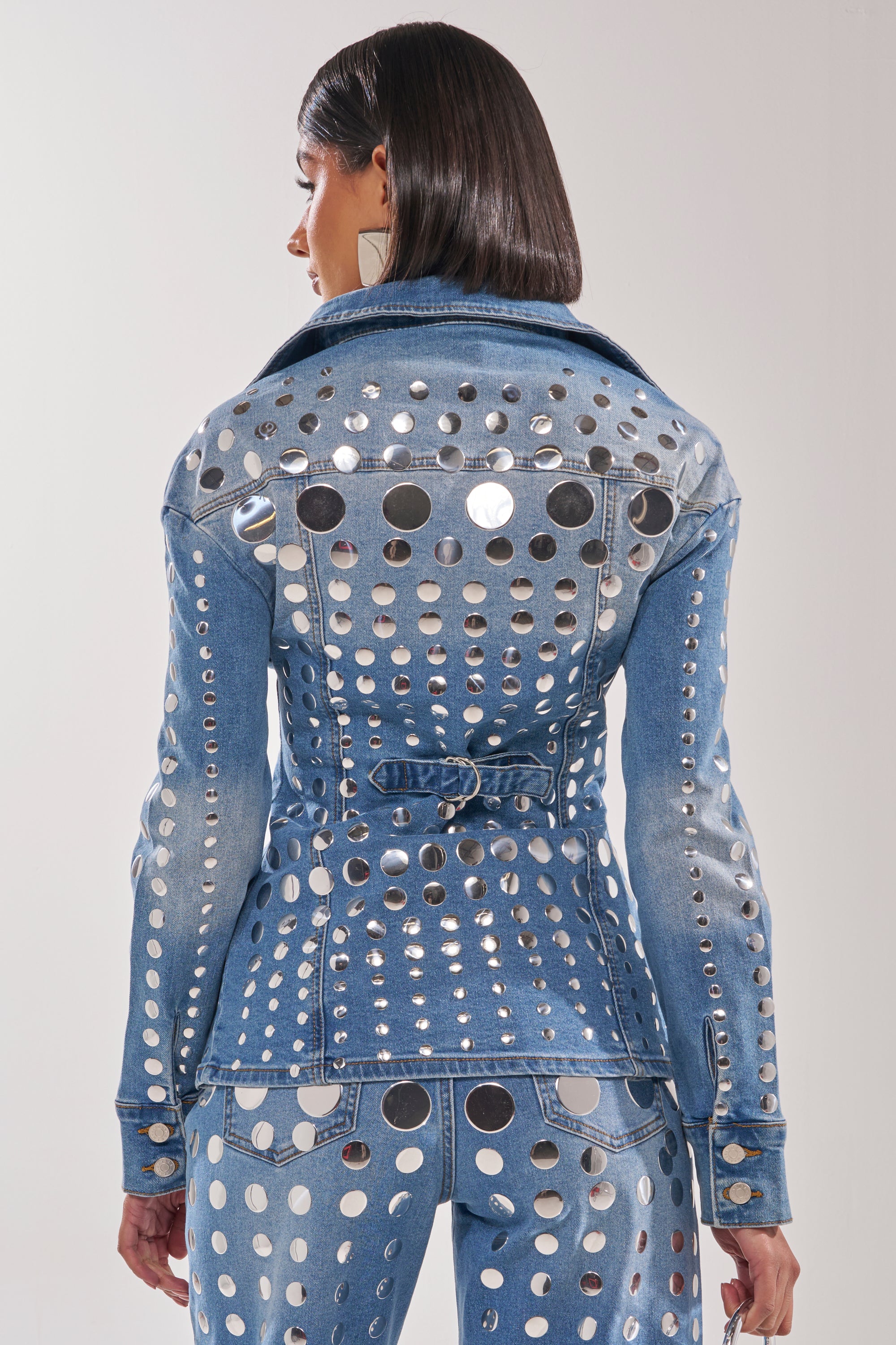 A woman with straight, dark hair faces away from the camera, showcasing the "STUDS EVERYWHERE BUTTON DOWN"—a waist-contouring denim top covered in metallic circular studs—against a plain background. 5