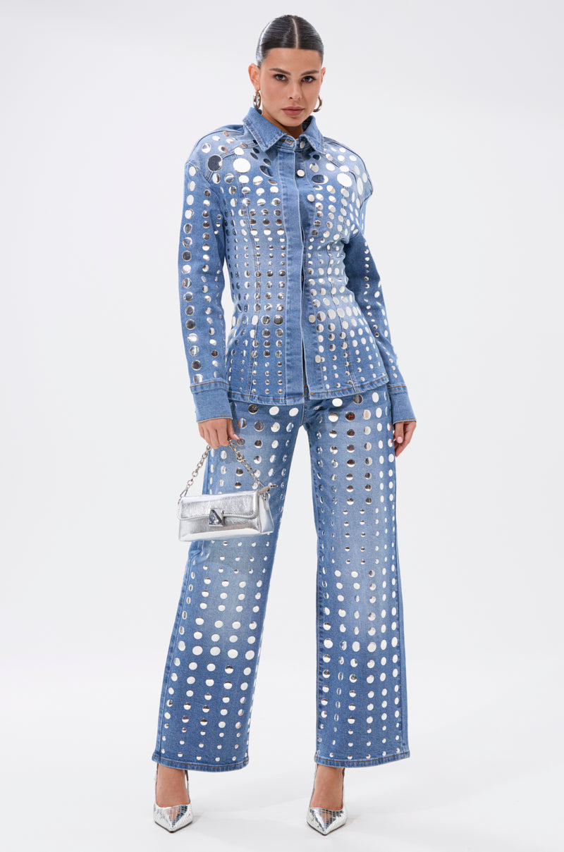 A woman poses against a plain white background in the STUDS EVERYWHERE BUTTON DOWN, a waist-contouring denim jacket with large silver grommets, paired with matching pants, a metallic purse, and silver pointed-toe heels.