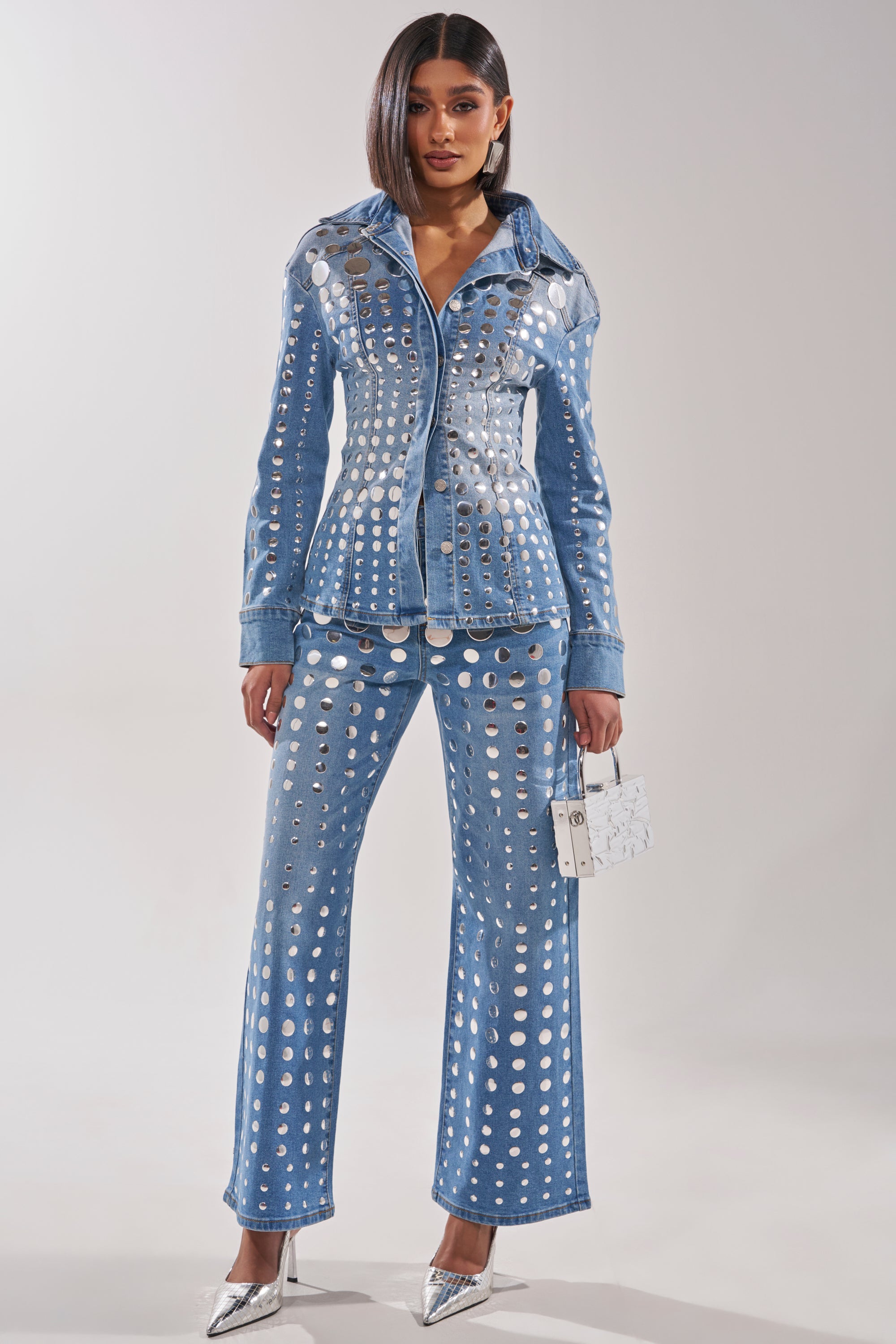 She wears the STUDS EVERYWHERE BUTTON DOWN, a studded denim top with matching pants featuring large silver grommets. The waist-contouring look is styled with a small white bag, pointed silver heels, and her sleek, straight bob. 6