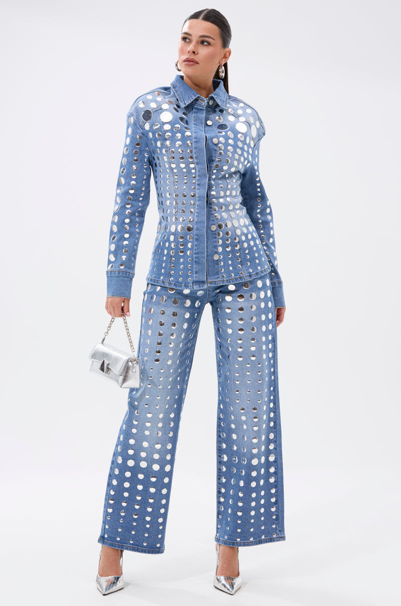 Wearing the STUDS EVERYWHERE BUTTON DOWN and matching denim pants, a woman stands confidently in a waist-contouring silhouette with metallic silver studs, holding a silver purse and wearing pointed-toe heels against a white background.