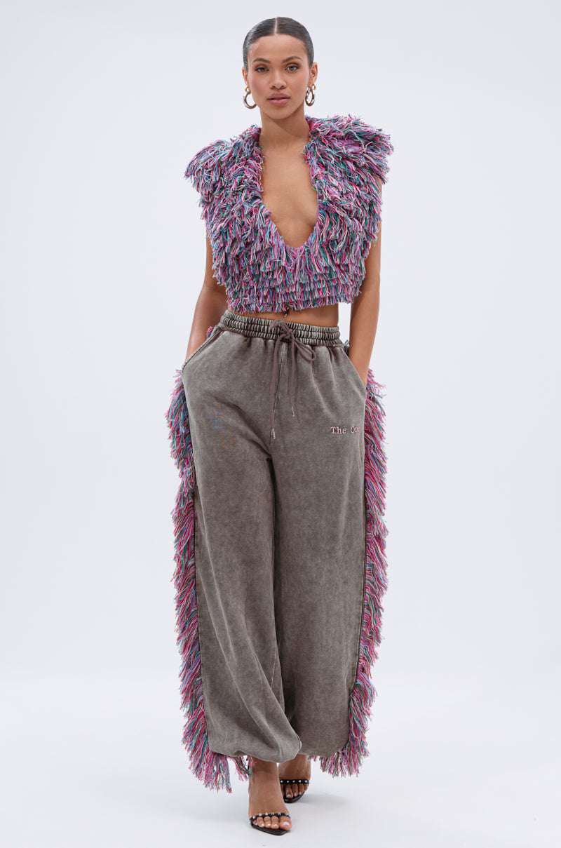 A woman stands against a plain background wearing THE ONE WIDE LEG JOGGER WITH FRINGE, featuring a textured, multicolored fringe top, loose gray wide-leg fit, paired with strappy heels and hoop earrings.