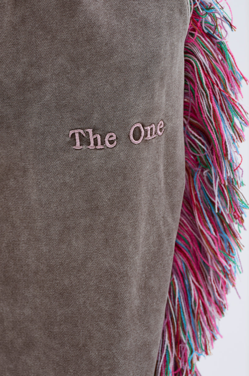 A close-up of brown fabric from THE ONE WIDE LEG JOGGER WITH FRINGE shows “The One” embroidered in pink, with colorful pink, green, and blue fringe along the edge of this unique jogger.