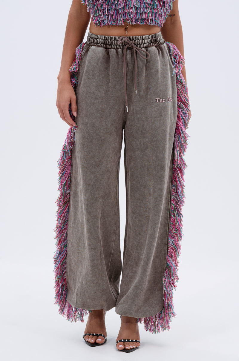 Someone wearing THE ONE WIDE LEG JOGGER WITH FRINGE, featuring multicolored fringe down the sides, styled with a matching fringed crop top and open-toe heels. Upper body is partially visible.