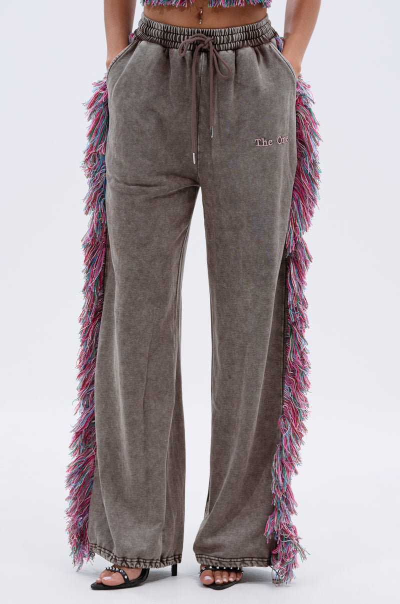 A person wears THE ONE WIDE LEG JOGGER WITH FRINGE in stone-washed gray, featuring colorful side fringe, a drawstring waist, and "The org" embroidery on one thigh, styled with open-toed heels.