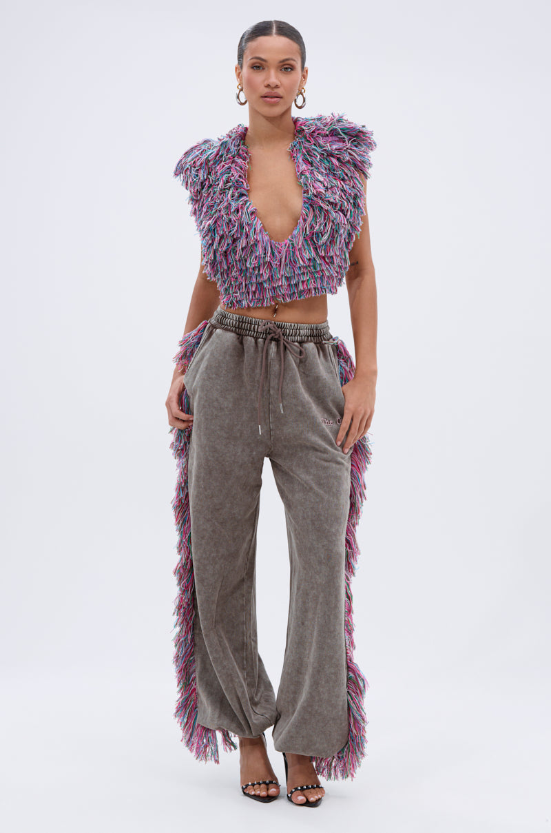 A woman stands facing forward, wearing THE ONE WIDE LEG JOGGER WITH FRINGE by AKIRA Label—gray high-waisted wide leg joggers featuring multicolored fringe details. She pairs them with hoop earrings and black heeled sandals.
