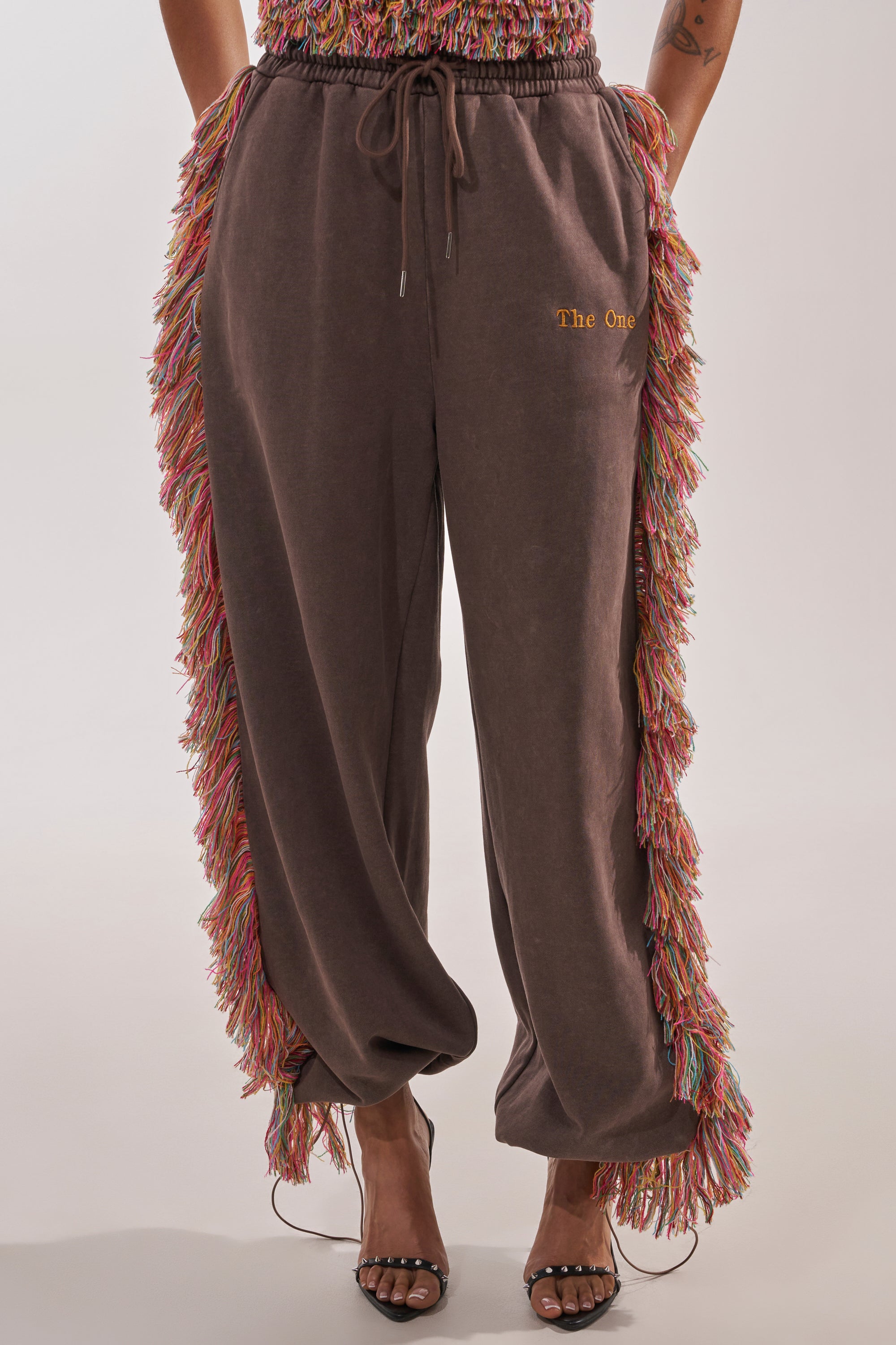 A person wears the THE ONE FRINGE JOGGER—a wide-leg brown pant with multicolored fringe along the side seams and "The One" embroidered in yellow on the upper thigh—paired with open-toe heels. 1