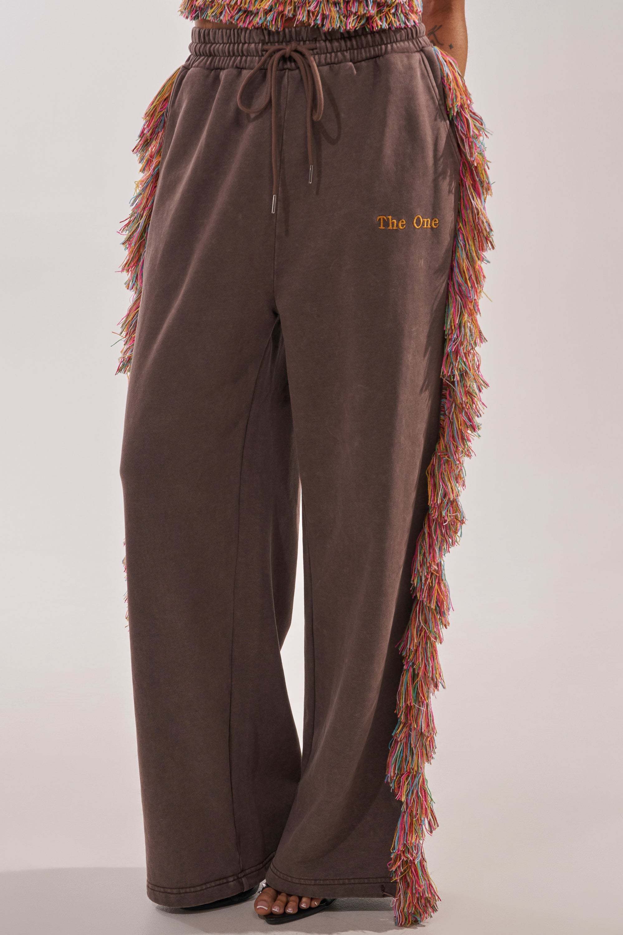The lower half of a person is shown wearing THE ONE FRINGE JOGGER: wide-leg brown sweatpants with colorful fringe along the outer sides and "The One" embroidered in yellow on the upper left thigh. 2