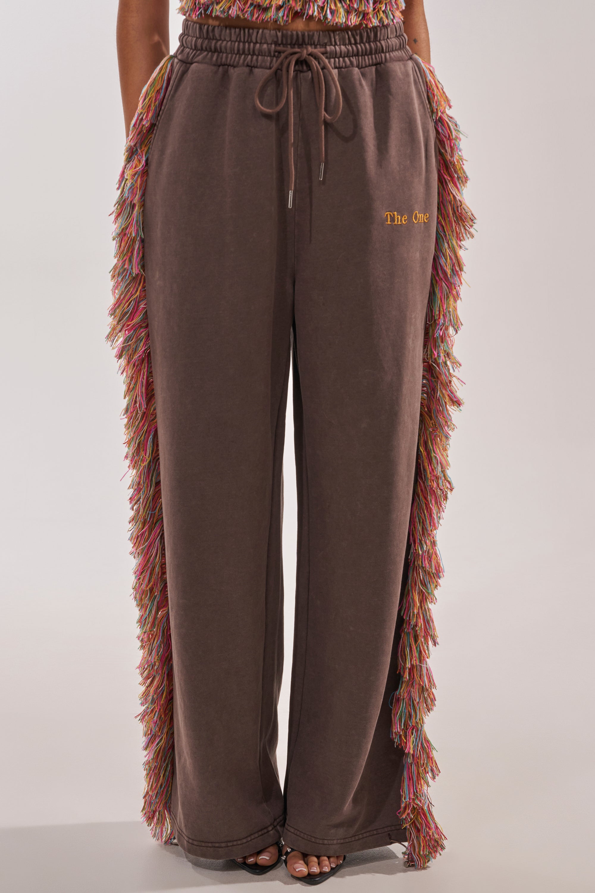 A person wearing THE ONE FRINGE JOGGER—wide-leg brown sweatpants with rainbow fringe on the side seams, a drawstring waist, and "The One" embroidered in yellow on the thigh; perfect for festivals. 3
