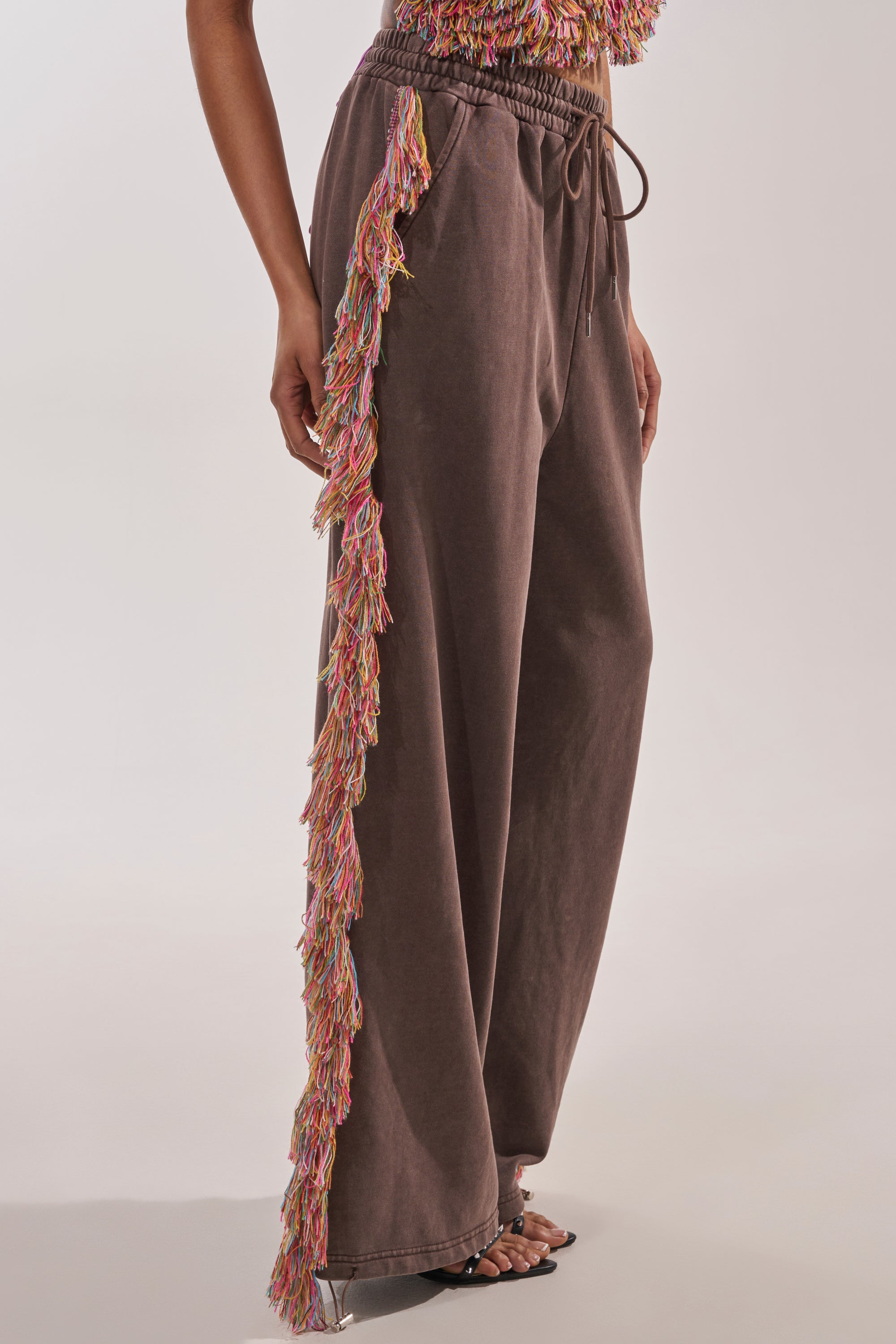 Wearing THE ONE FRINGE JOGGER, featuring wide-leg brown sweatpants with colorful side fringe, paired with heeled sandals. Only the lower half is shown, spotlighting this unique festival streetwear piece. 4