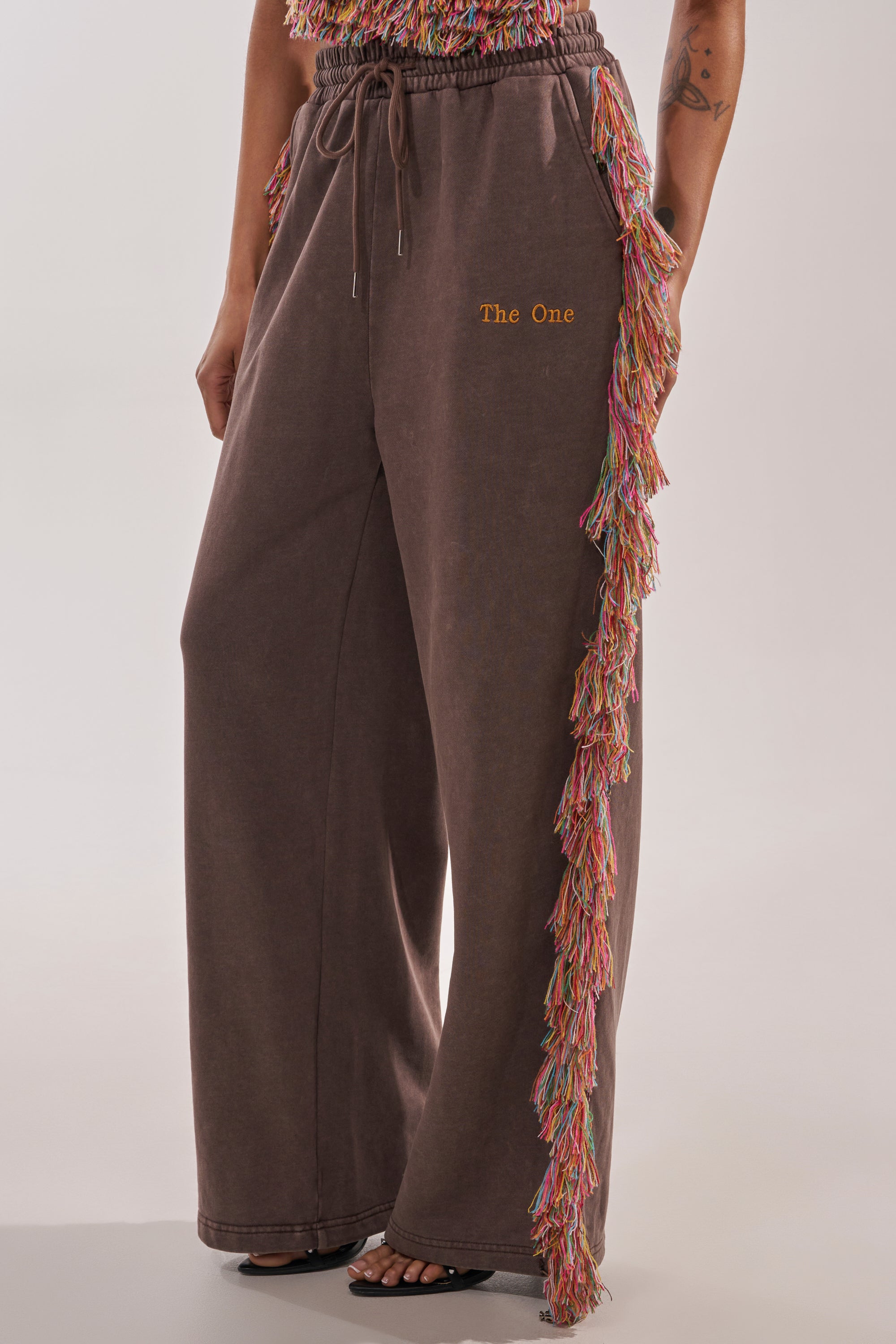 Someone wears THE ONE FRINGE JOGGER—wide-leg brown sweatpants with a drawstring waist, multicolored fringe down one leg, and "The One" embroidered in yellow on the left thigh. Only the lower half is shown; perfect for festival streetwear. 5