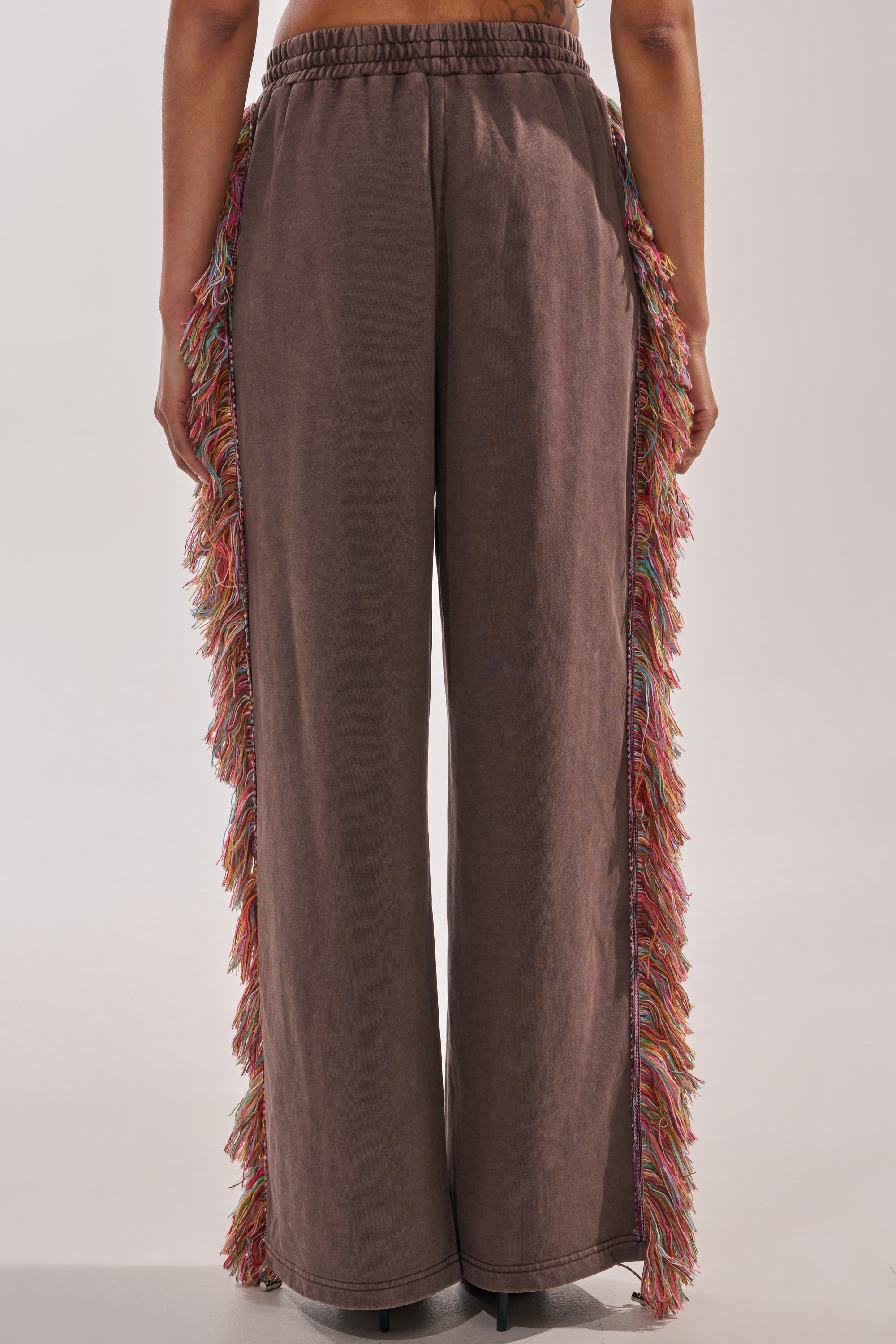 Photographed from behind against a plain background, a person wears THE ONE FRINGE JOGGER—wide-leg brown sweatpants featuring colorful fringe along the outer seams, ideal for festival streetwear. 6