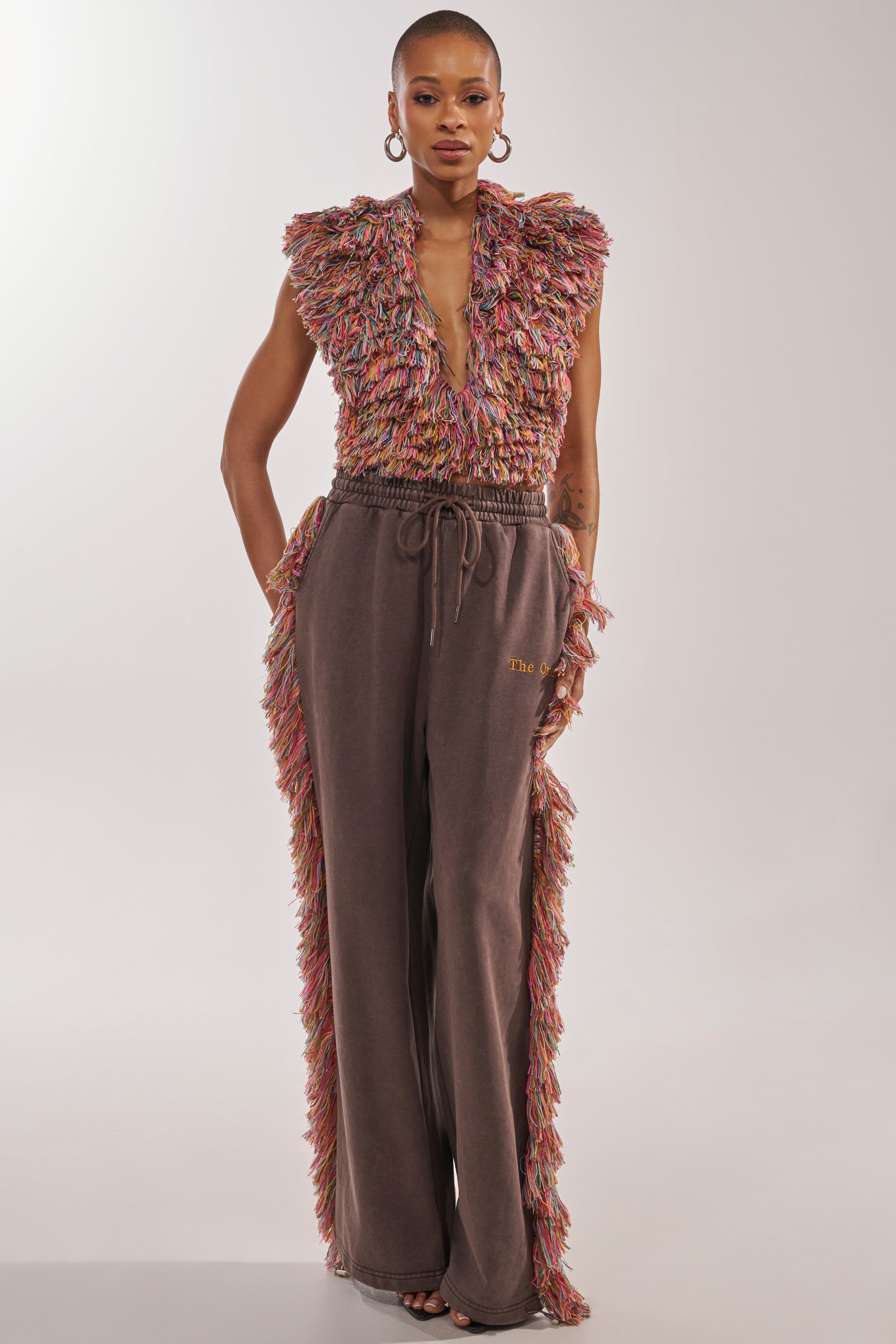 A person faces forward in THE ONE FRINGE JOGGER, a wide-leg brown pant with multicolored fringe down the sides, styled with a matching textured fringed top for a striking festival streetwear look against a plain light background. 7